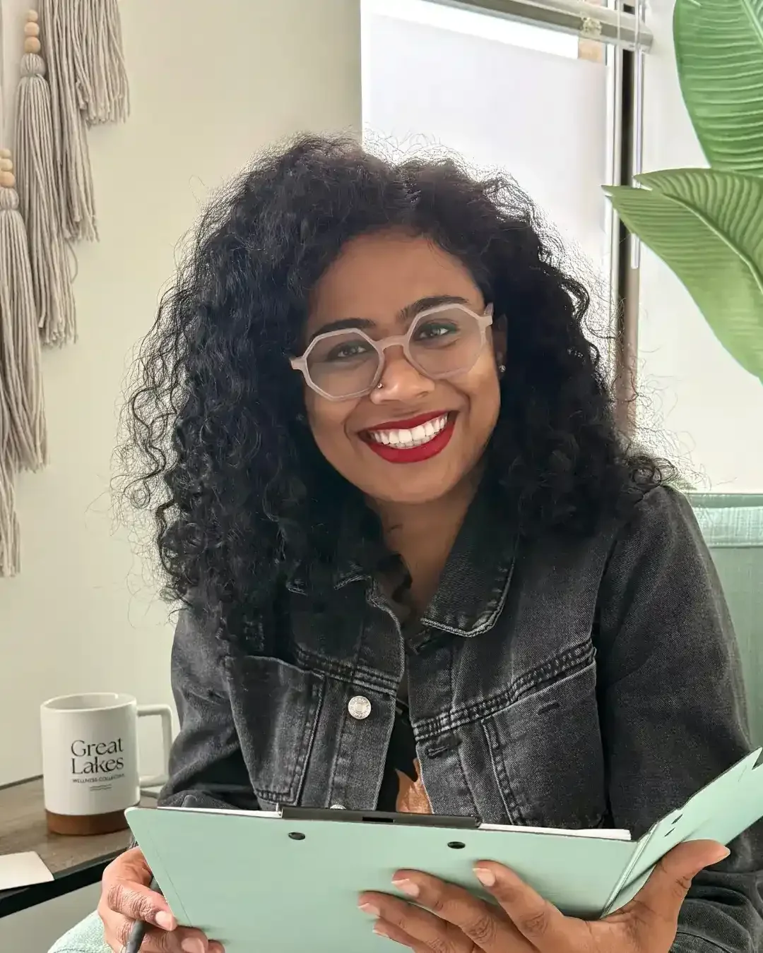 Ashish | Smiling woman with curly hair and glasses holding a notebook, sitting near a window with green plants.