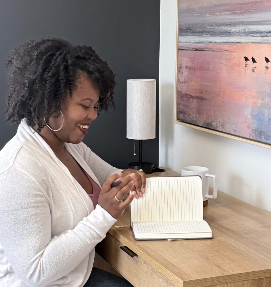 A woman smiling and writing in a notebook at a wooden desk next to a lamp and a mug.