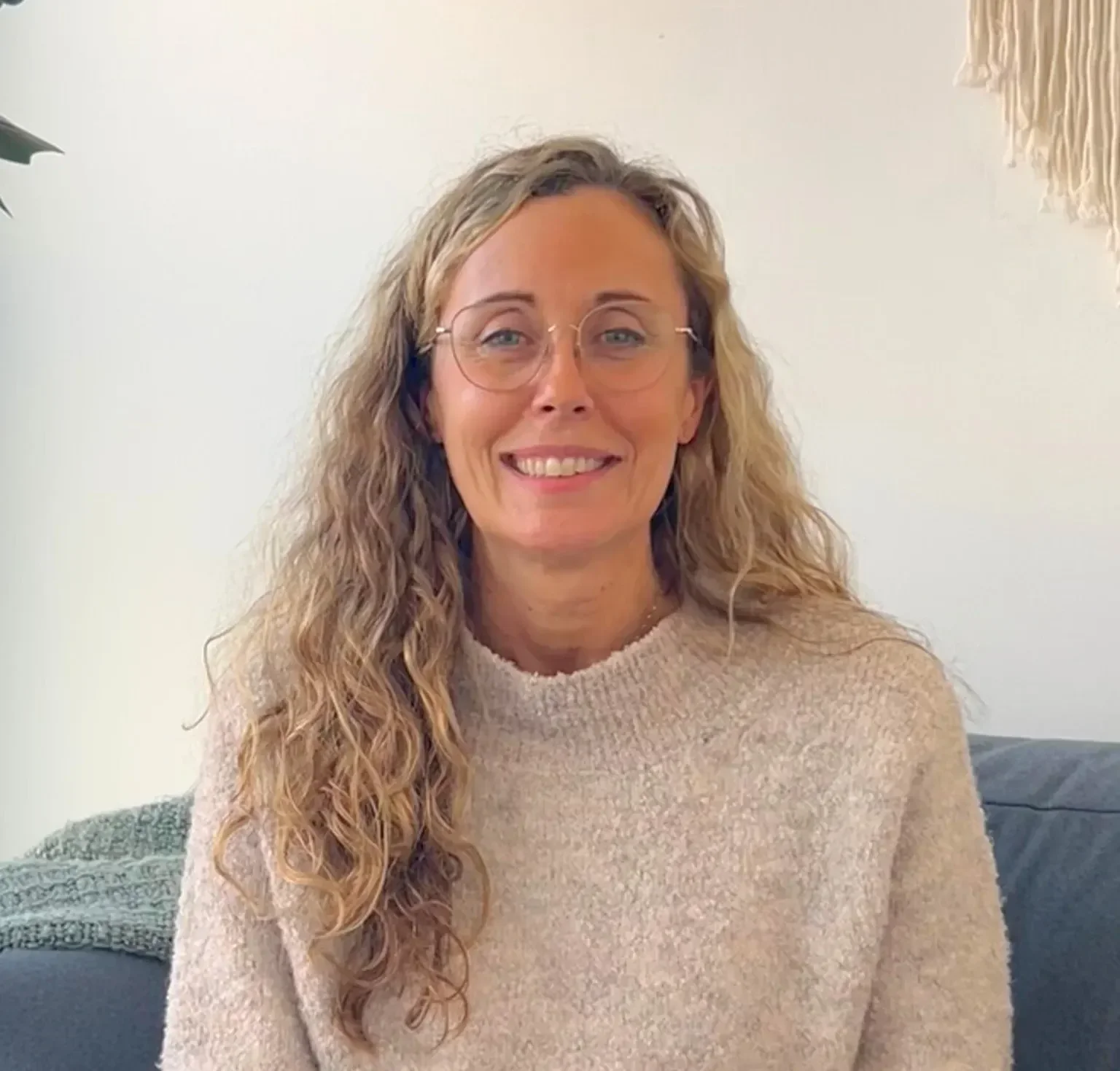 Sandra | Social Worker | A woman with long, curly blonde hair and glasses smiling while sitting on a couch in a cozy room.