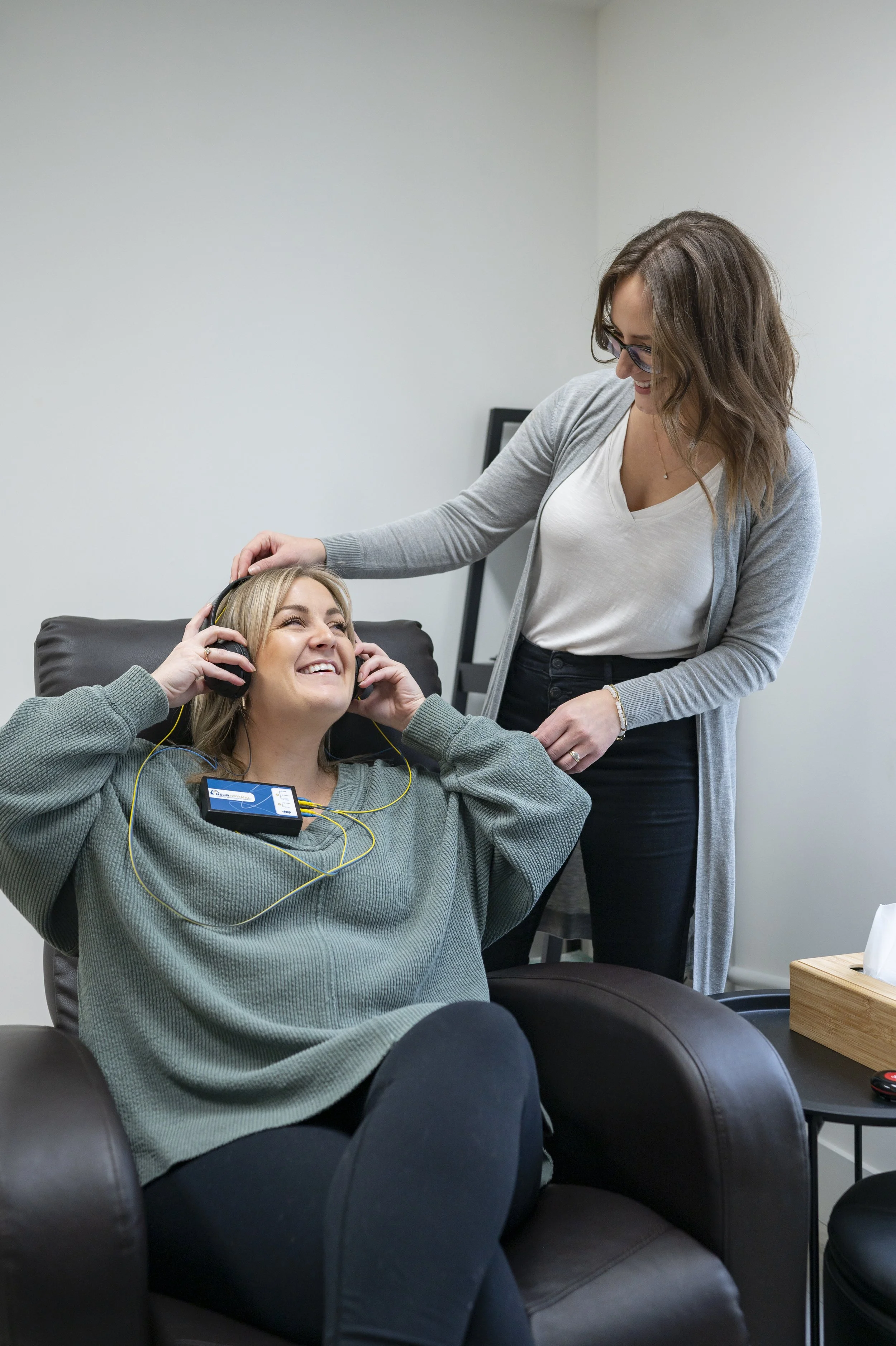 A woman sitting in a black leather chair, smiling and listening to music with headphones, while another woman standing beside her, gently touching her head and smiling, in a well-lit room with a minimalist decor.