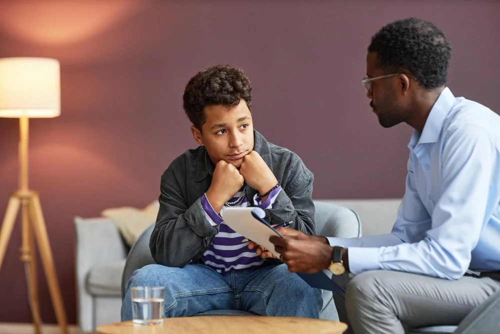 A young boy sitting in a therapist's office, listening attentively while a therapist talks to him, with a glass of water on the table and a lamp in the background.