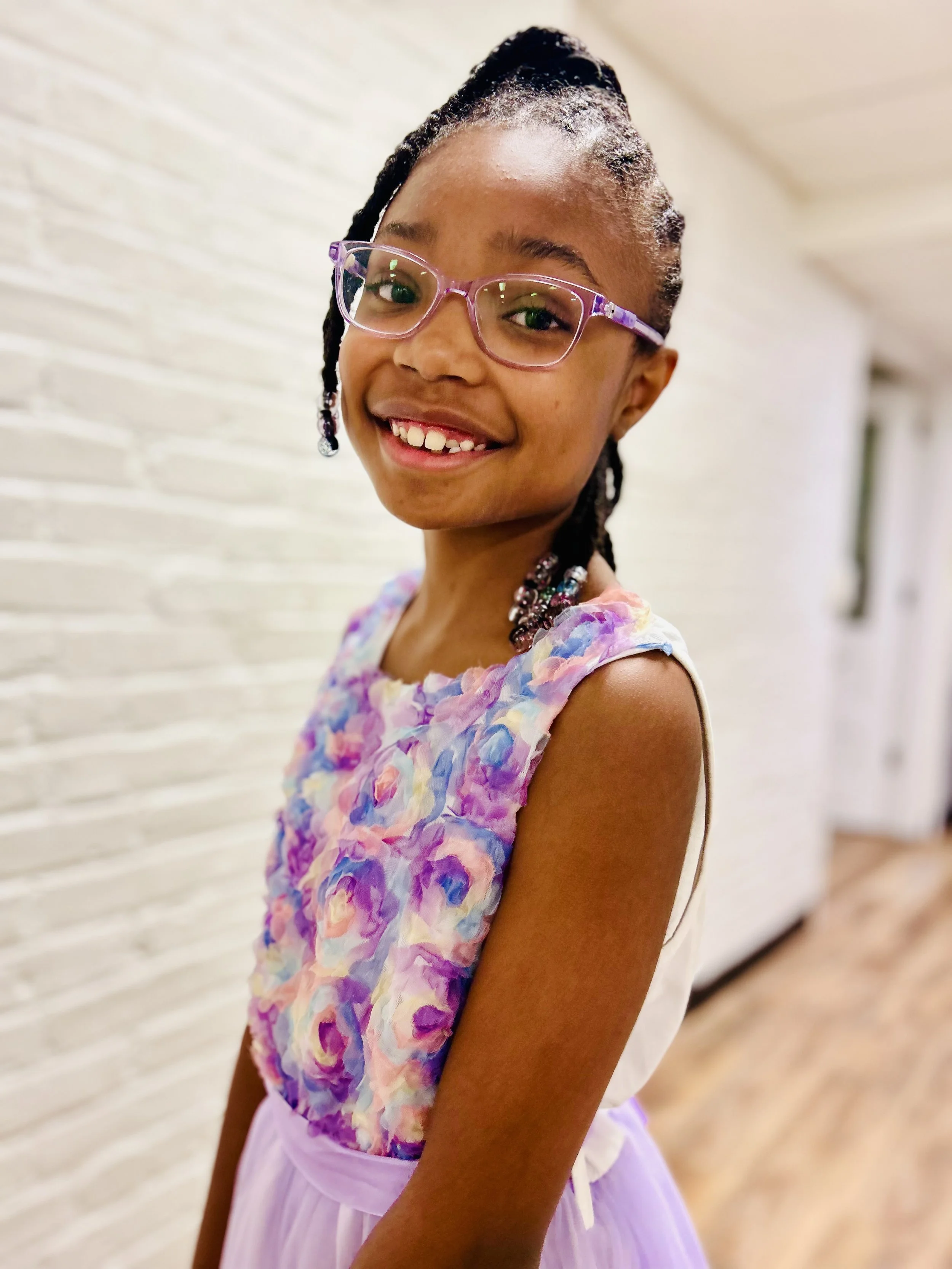 Smiling young girl with glasses and beaded hair, wearing a floral dress, standing indoors against a light-colored brick wall.