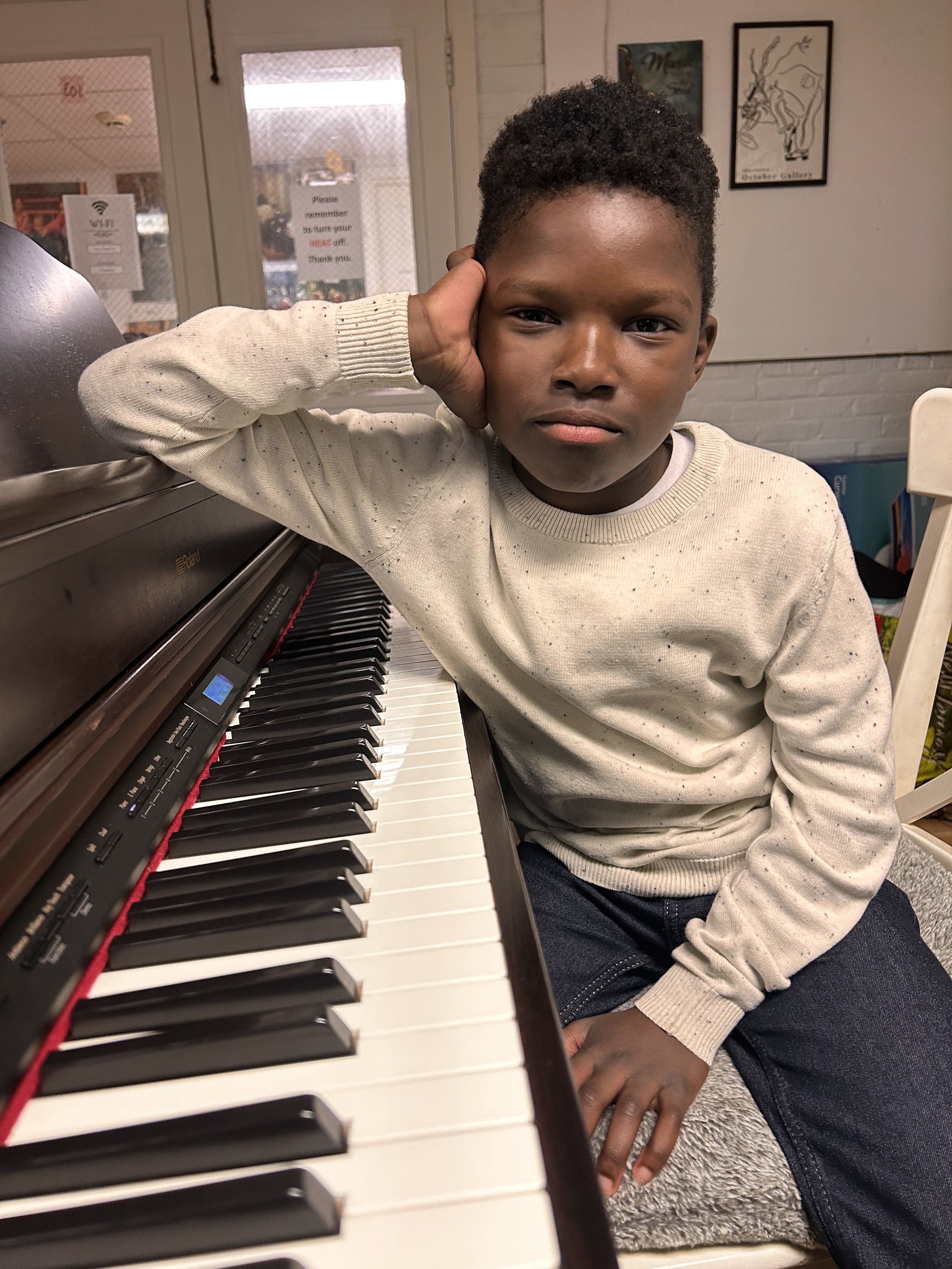 A young boy sitting at a piano, resting his head on his left hand, looking directly at the camera with a neutral expression.