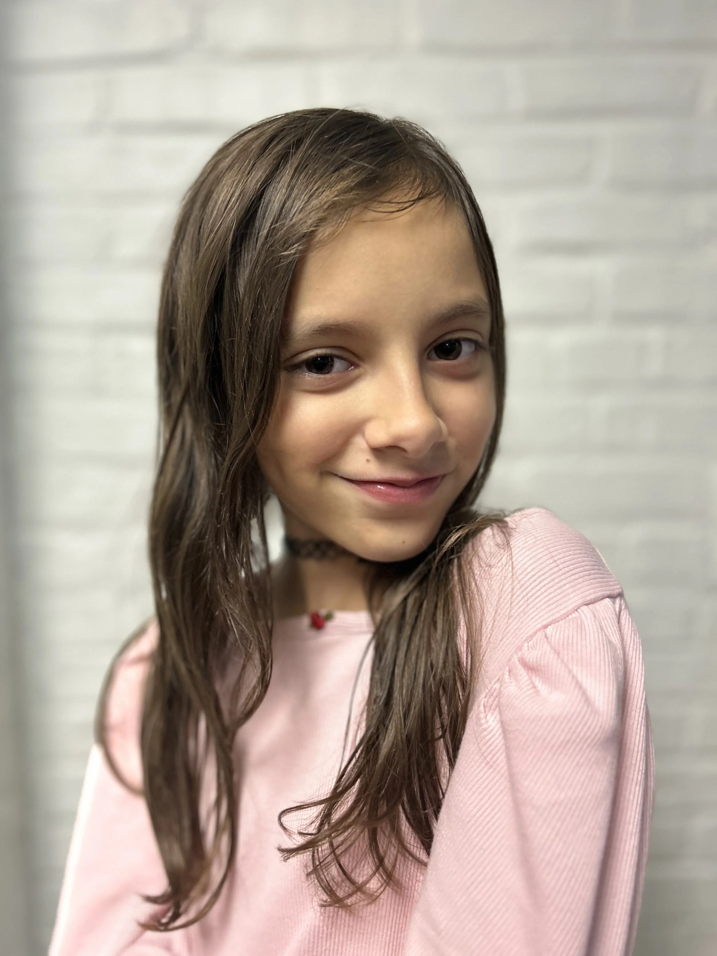 Smiling young girl with long brown hair wearing a pink shirt and a black choker necklace, standing against a white brick wall.