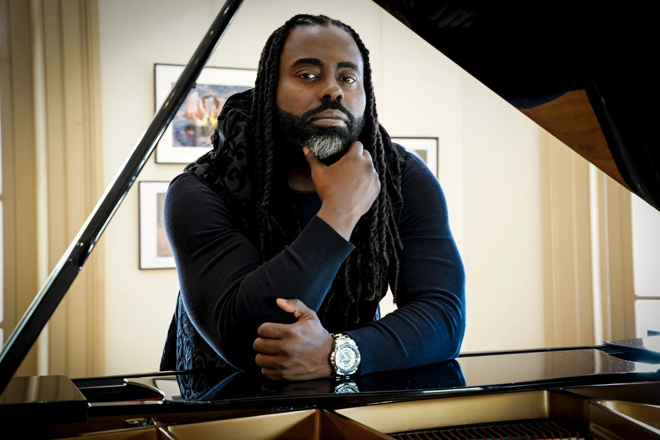 A man with long dreadlocks, beard, and dark skin posing with his hand on his chin, sitting behind a grand piano.