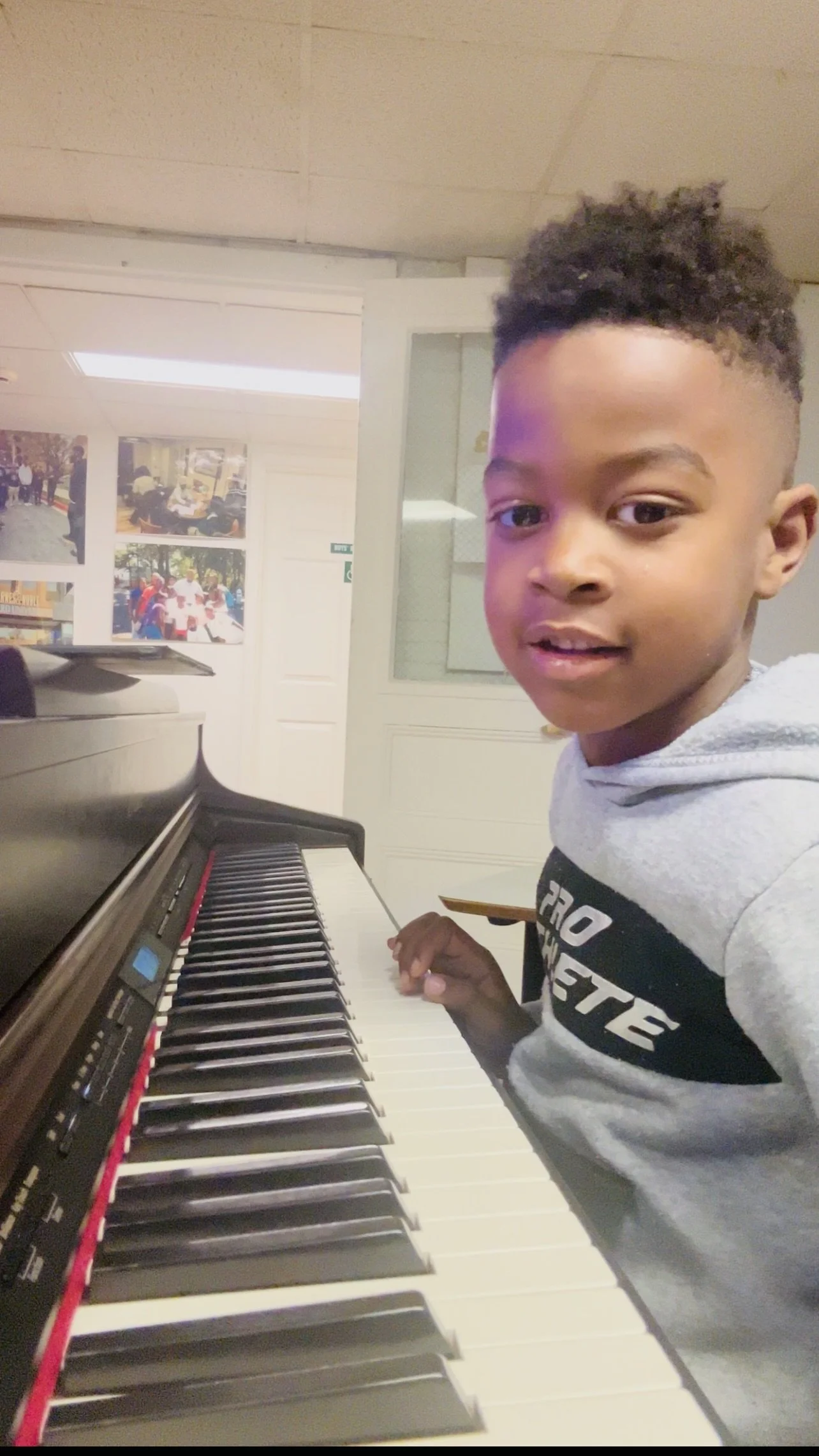 A young boy with short curly hair playing a piano in a room with photo posters on the wall.
