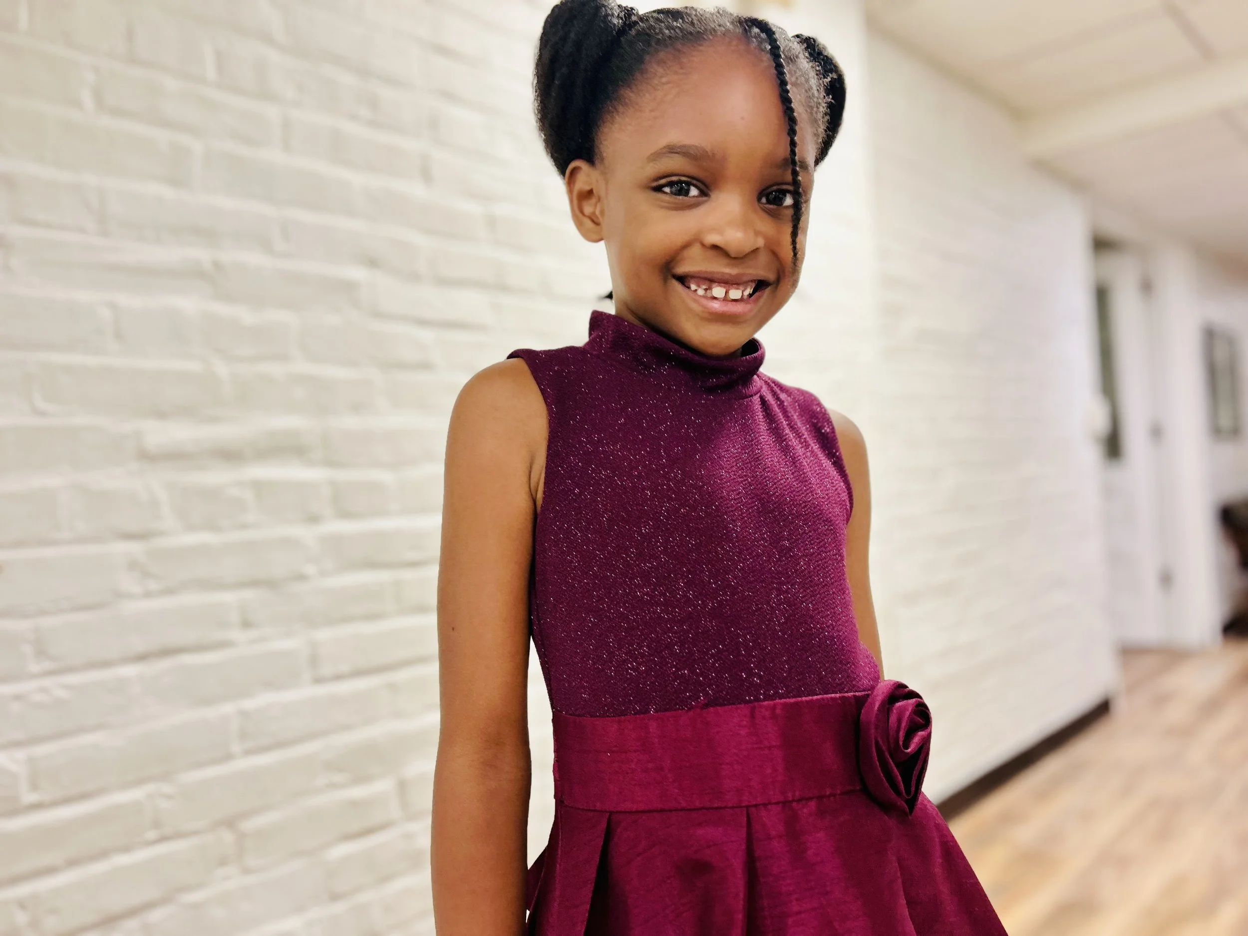 A vocal Student  with braided hair, smiling and wearing a sleeveless maroon dress with a bow at the waist, standing in front of a white brick wall.