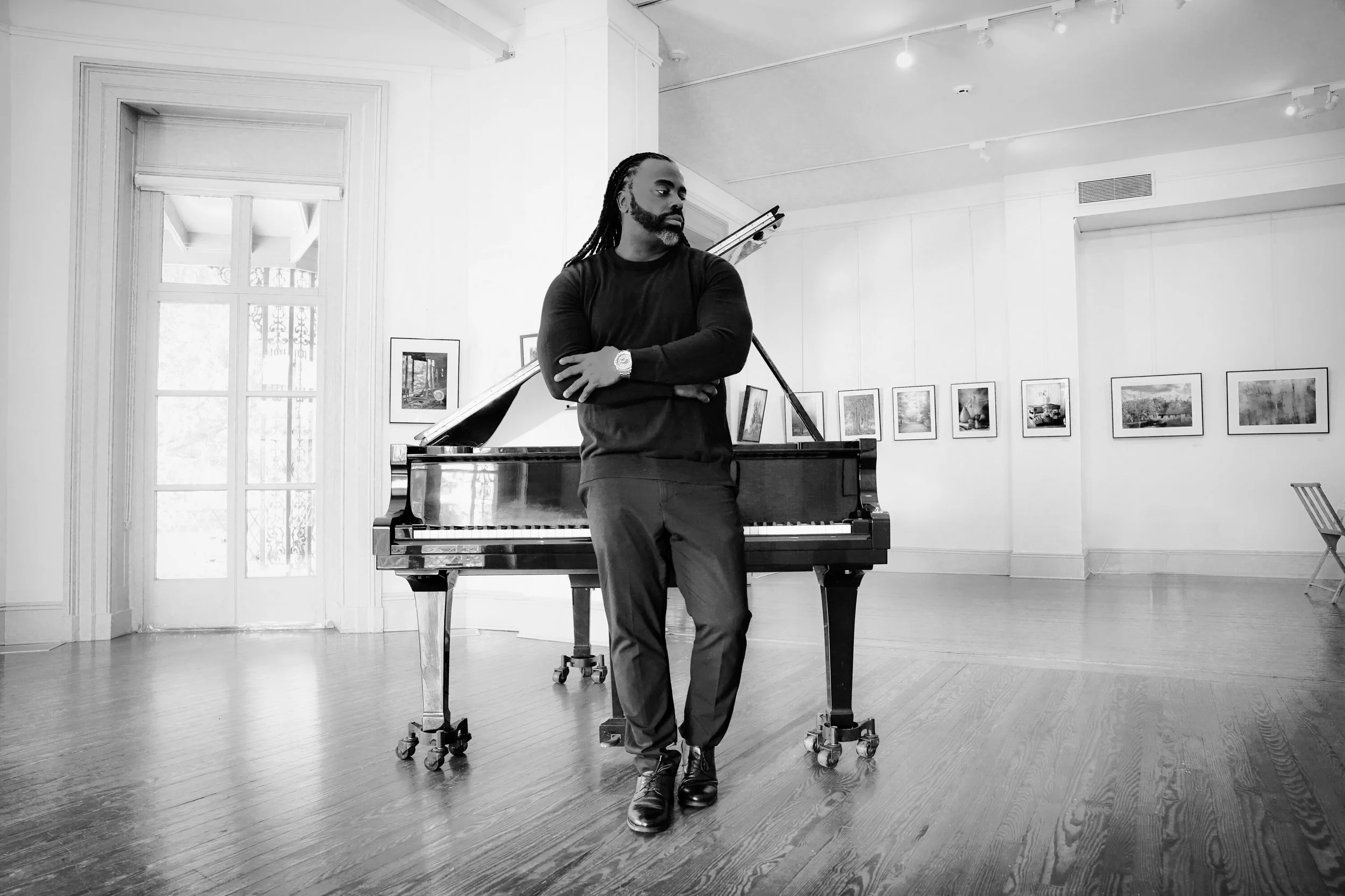 A man with long dreadlocks and a beard standing crosses arms in front of a grand piano inside an art gallery with framed pictures on the white walls.