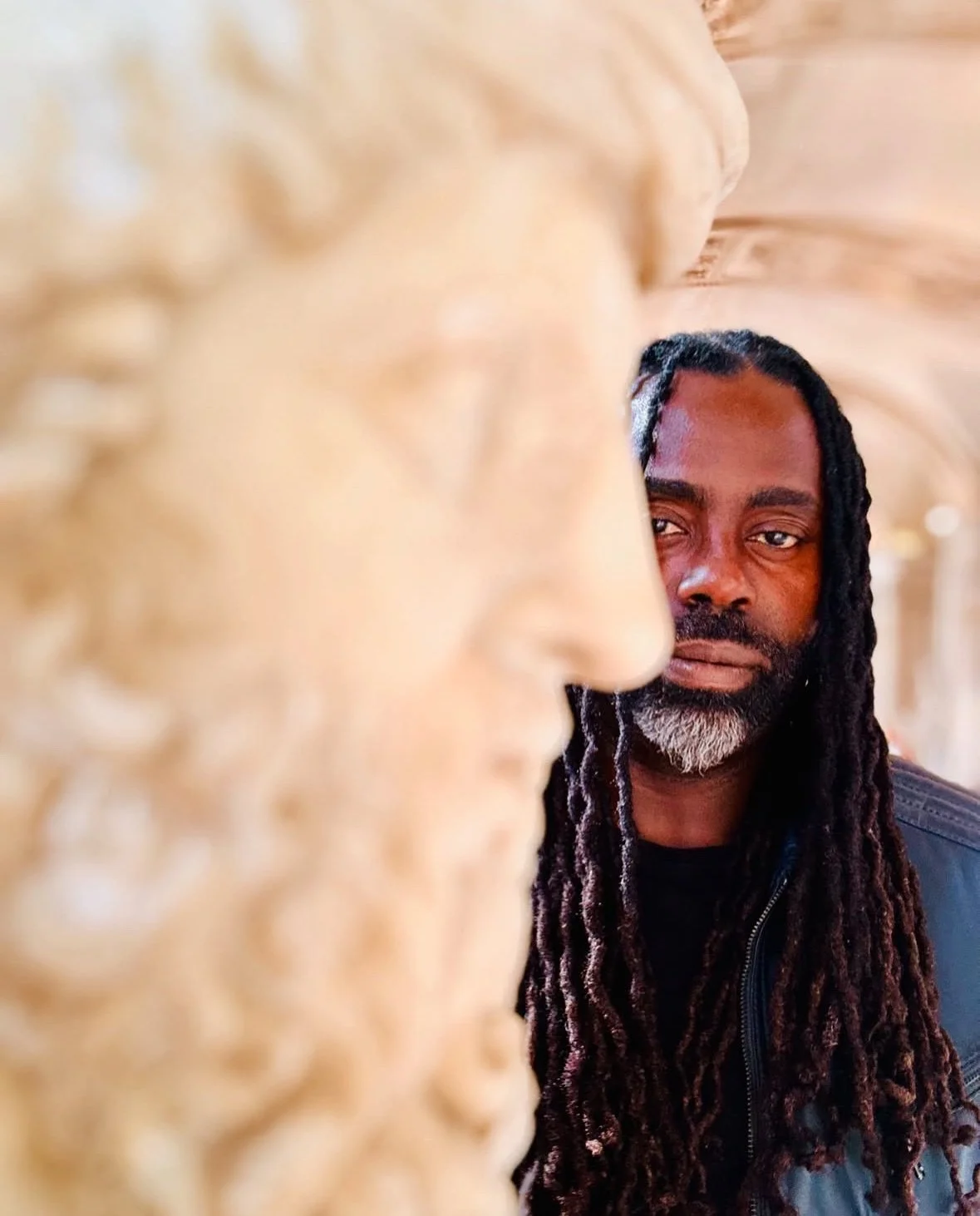 A man with long dreadlocks and a beard looking at a golden statue sculpture.