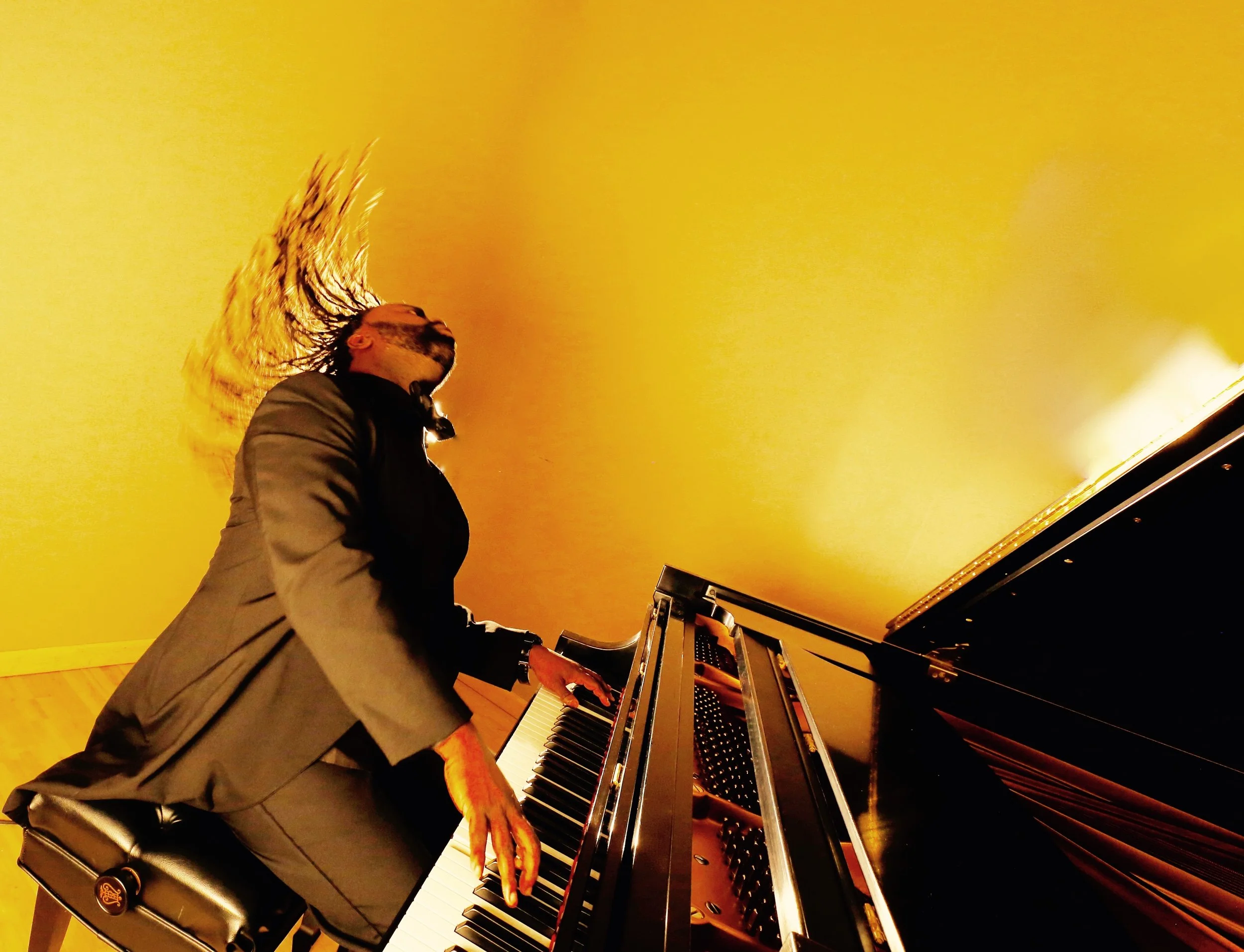 A person playing a grand piano with a yellow backdrop, head tilted back, hair flying, wearing glasses and a gray suit.