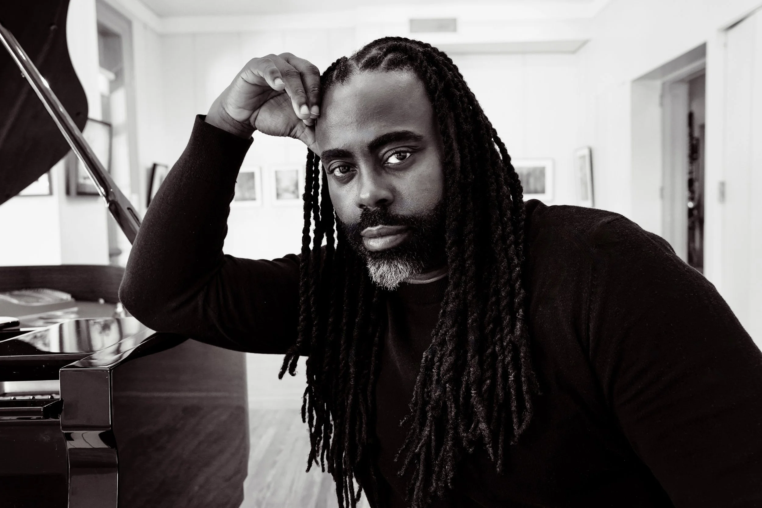 Portrait of a man with long dreadlocks and a beard, sitting beside a piano in a room with framed photos on the wall.