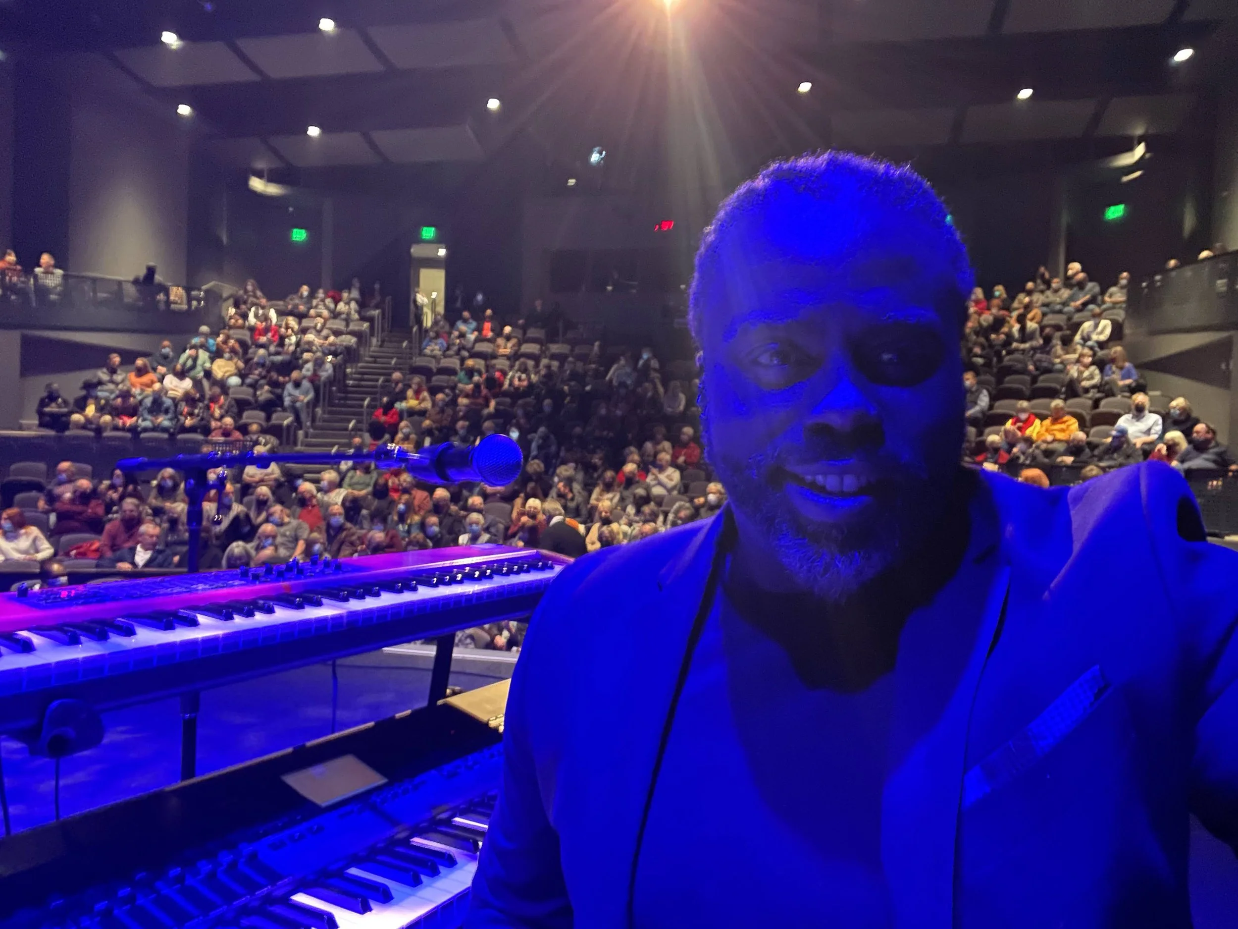 A man taking a selfie on stage in front of a grand piano and keyboard, in a theater with an audience seated, illuminated by purple and blue stage lighting.