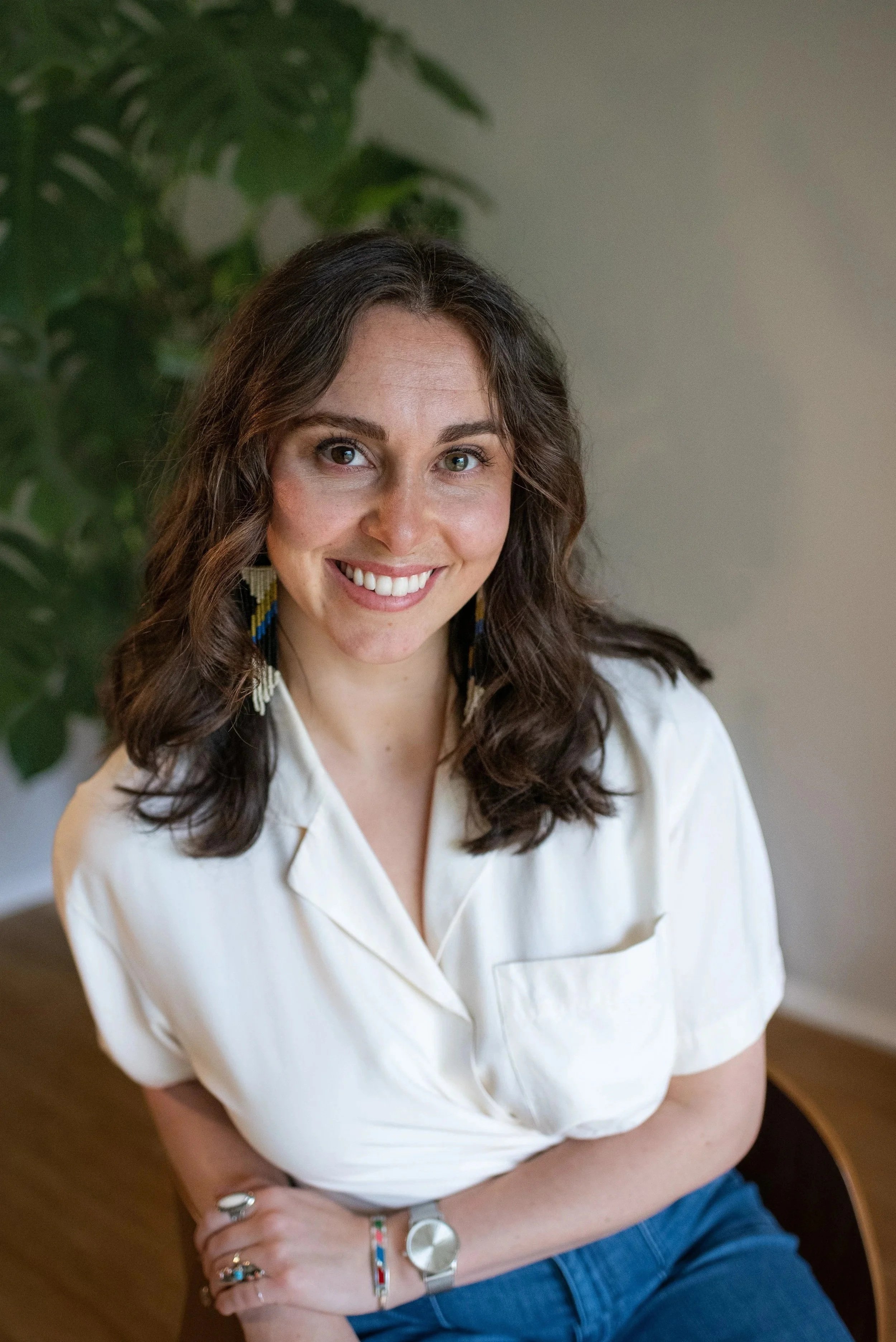 A woman with wavy brown hair and blue eyes, smiling, wearing a white shirt, colorful earrings, a watch, and several rings, sitting indoors with a large plant in the background.