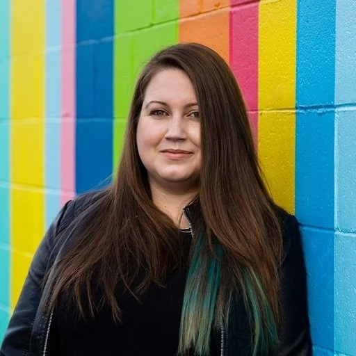 Woman with long brown hair and blue-green tips, standing against a multicolored painted brick wall.
