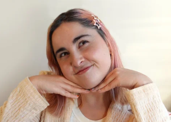 A young woman with pink hair and a hair clip, smiling with her hands under her chin in a playful pose.