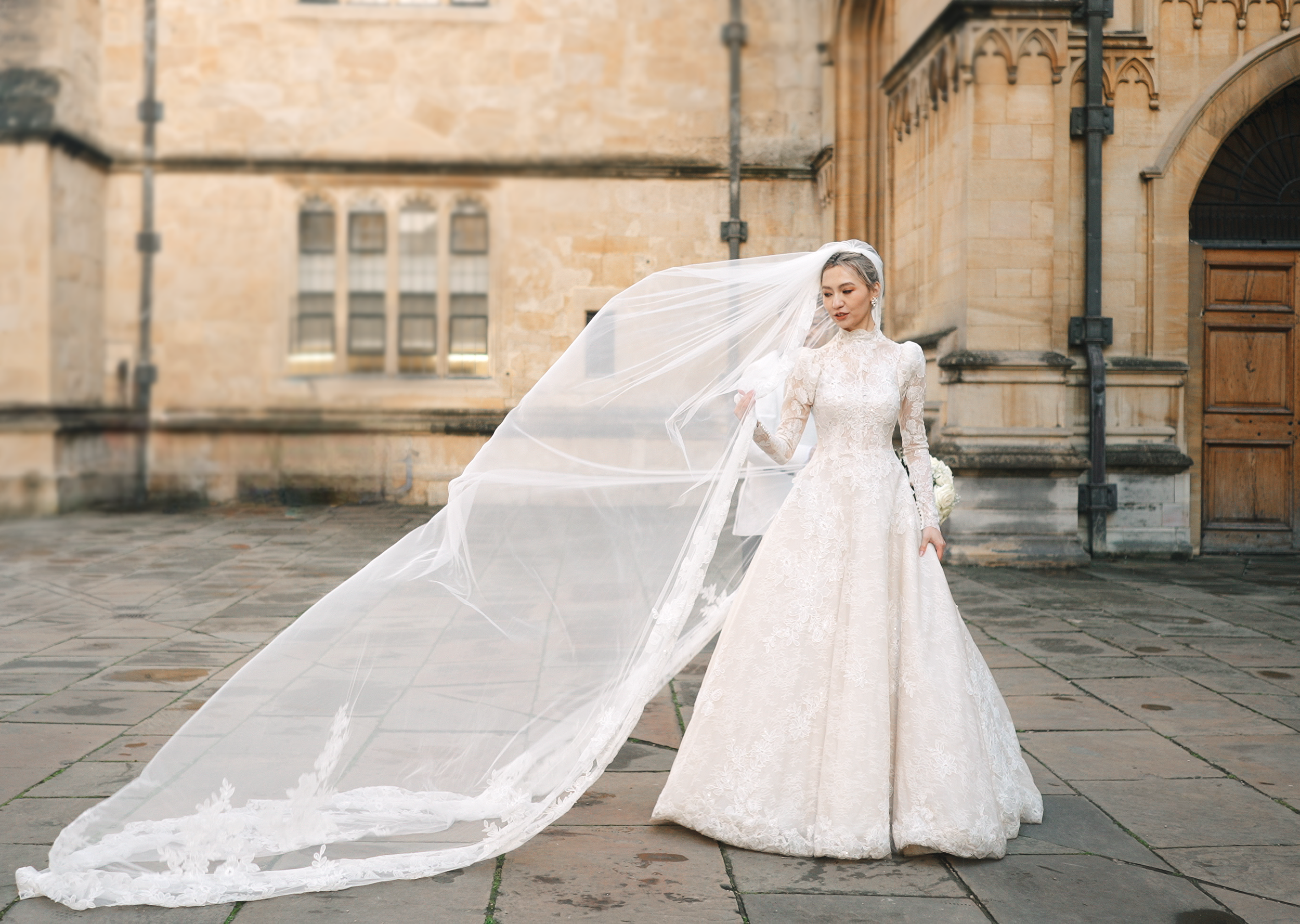 A bride in a long lace wedding gown with a high neckline and long sleeves, standing outdoors on a stone patio in front of a historic stone building with wooden doors and multiple windows. She is holding the edge of her long veil which billows in the wind, and she has short gray hair and earrings.