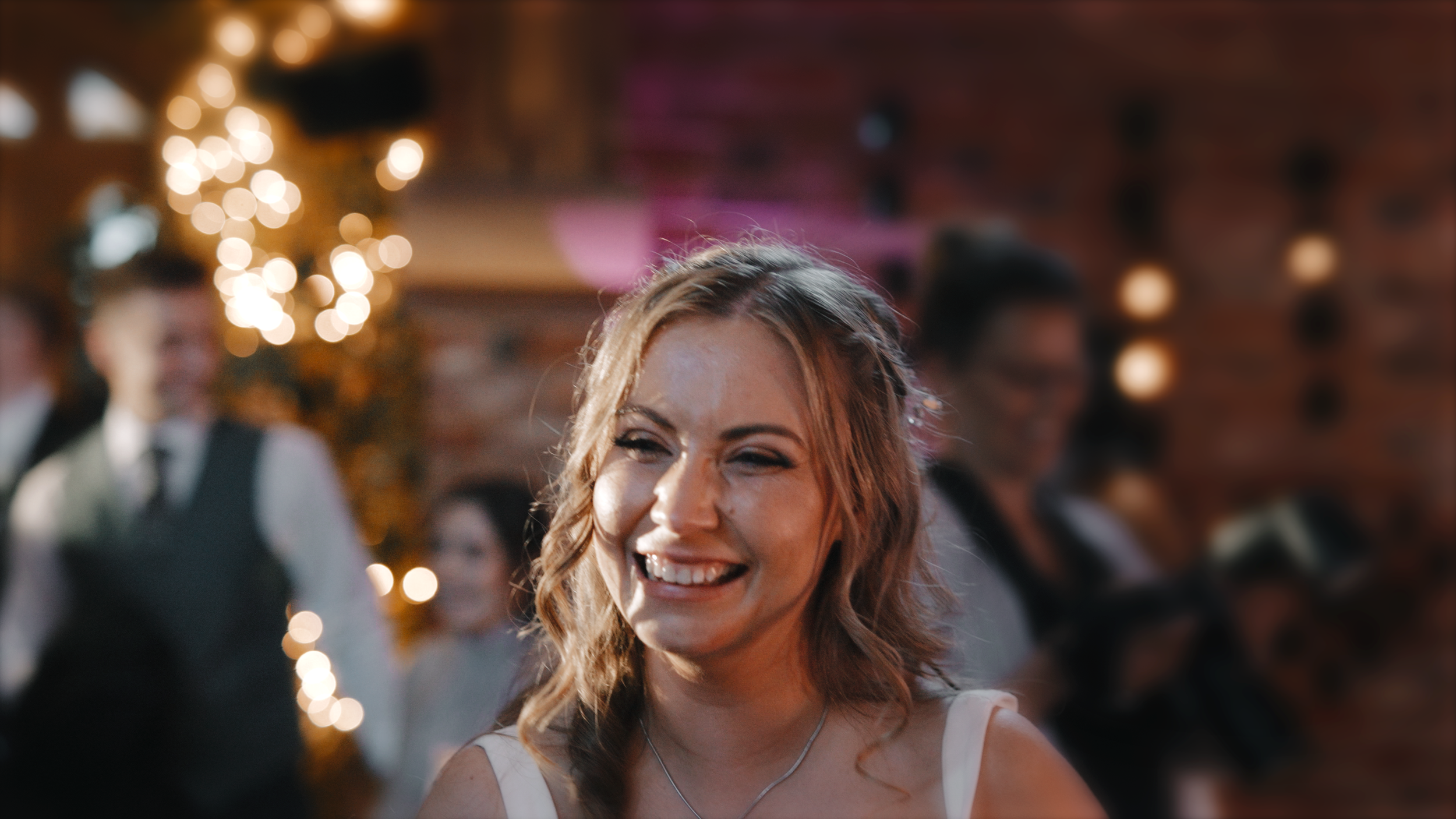 A woman with light brown, wavy hair, smiling at a festive gathering with blurred background lights and people.