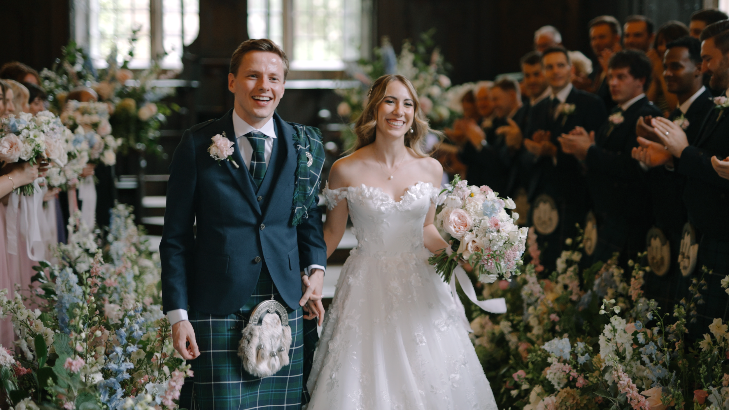 A bride and groom holding hands and smiling as they walk down the aisle during their wedding ceremony, surrounded by guests and floral arrangements.