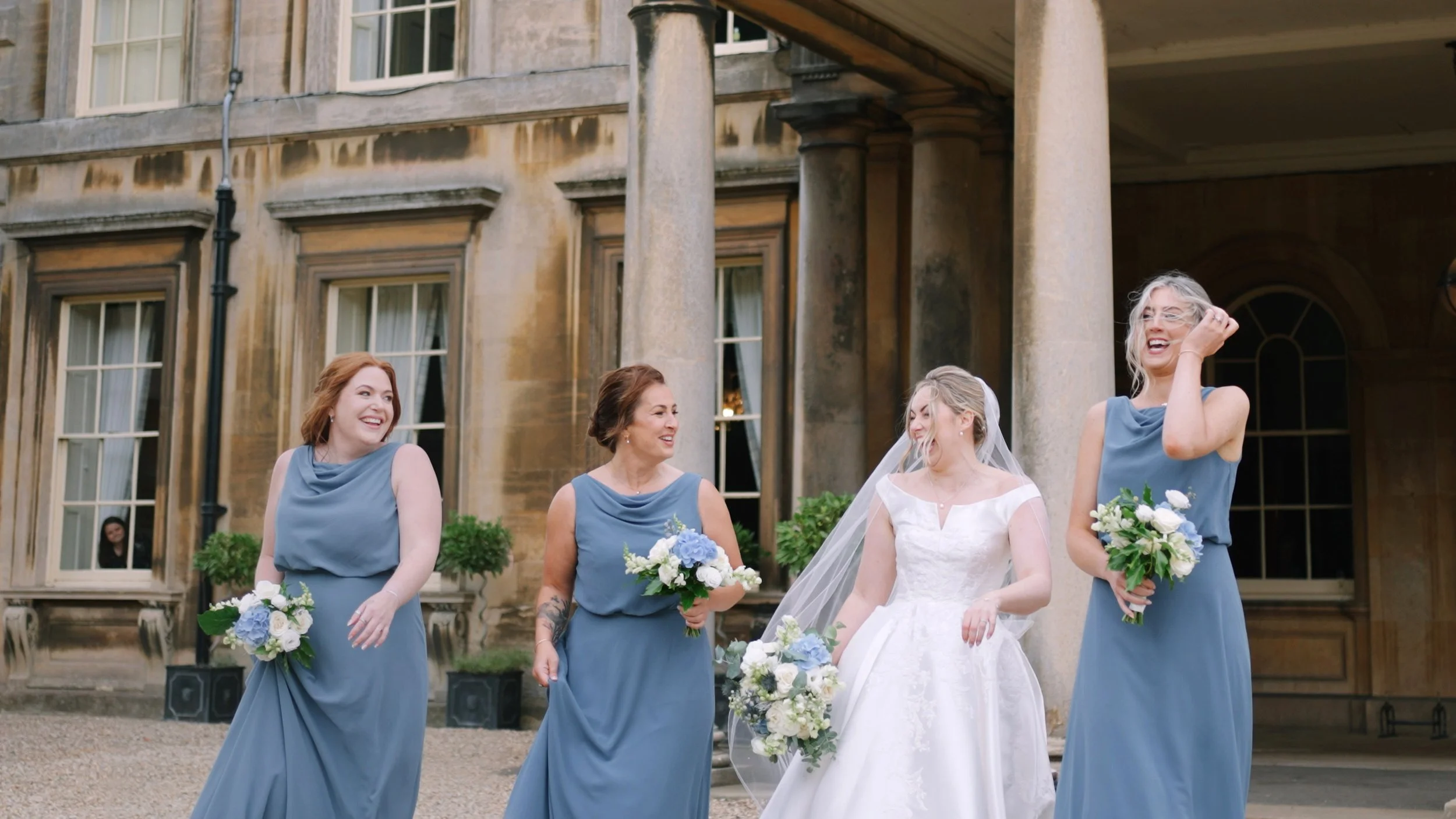 A bride in a white gown and veiled headpiece laughing with four bridesmaids in blue dresses holding bouquets, standing outside an elegant historic building with large windows and stone pillars.