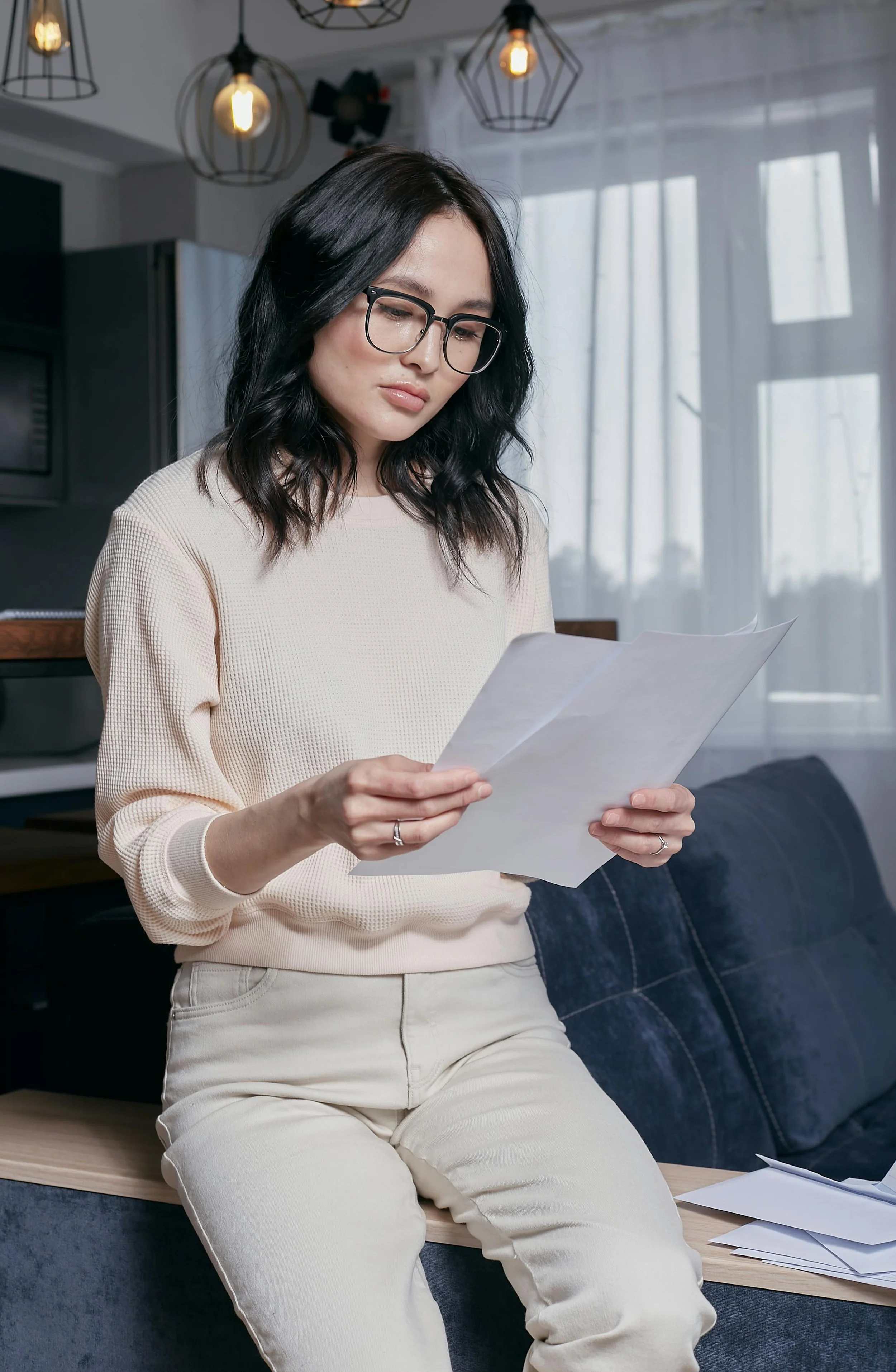 A woman sitting on a bench in a modern kitchen, reading a paper. She has black wavy hair, wears glasses, a cream sweater, and light-colored pants. There are paper sheets on the bench and sunlight coming through a window with white curtains.