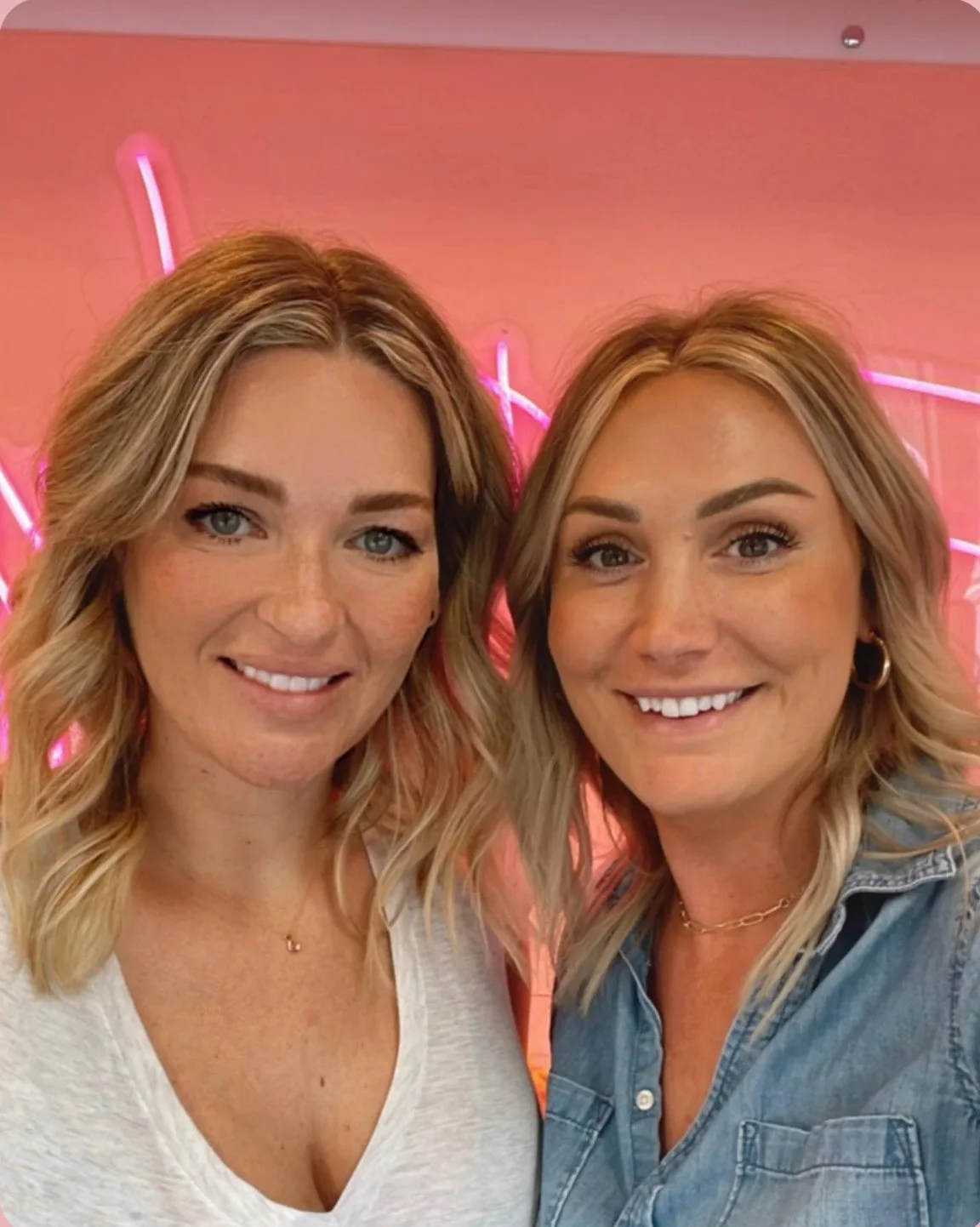 Two women smiling in front of a pink neon background.