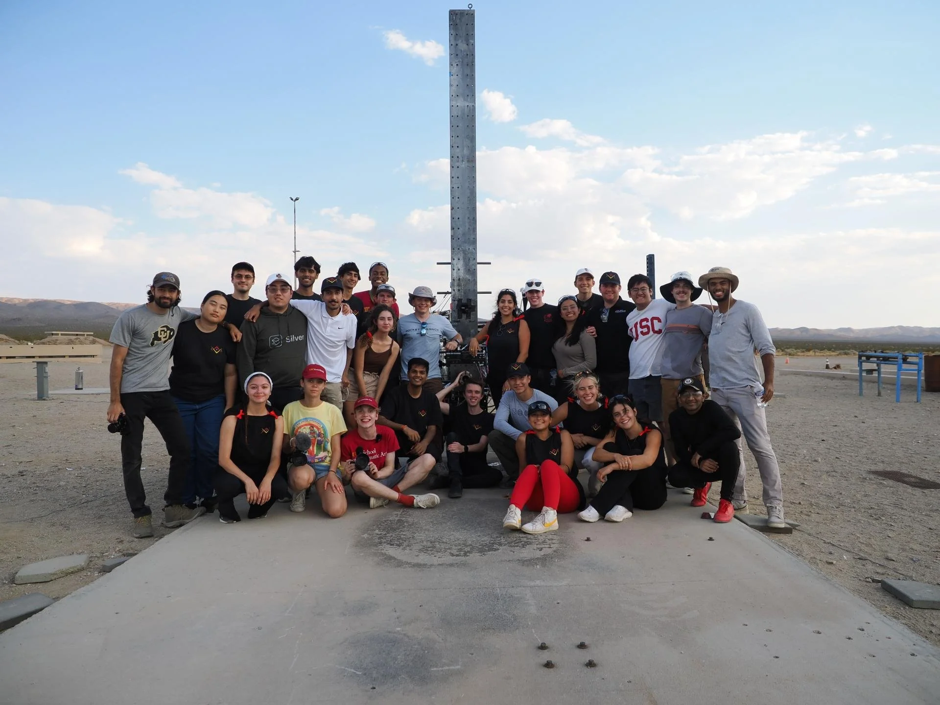 Group of people gathered outdoors in a desert, standing and sitting in front of a tall metal structure with holes, possibly an aerospace or scientific installation.