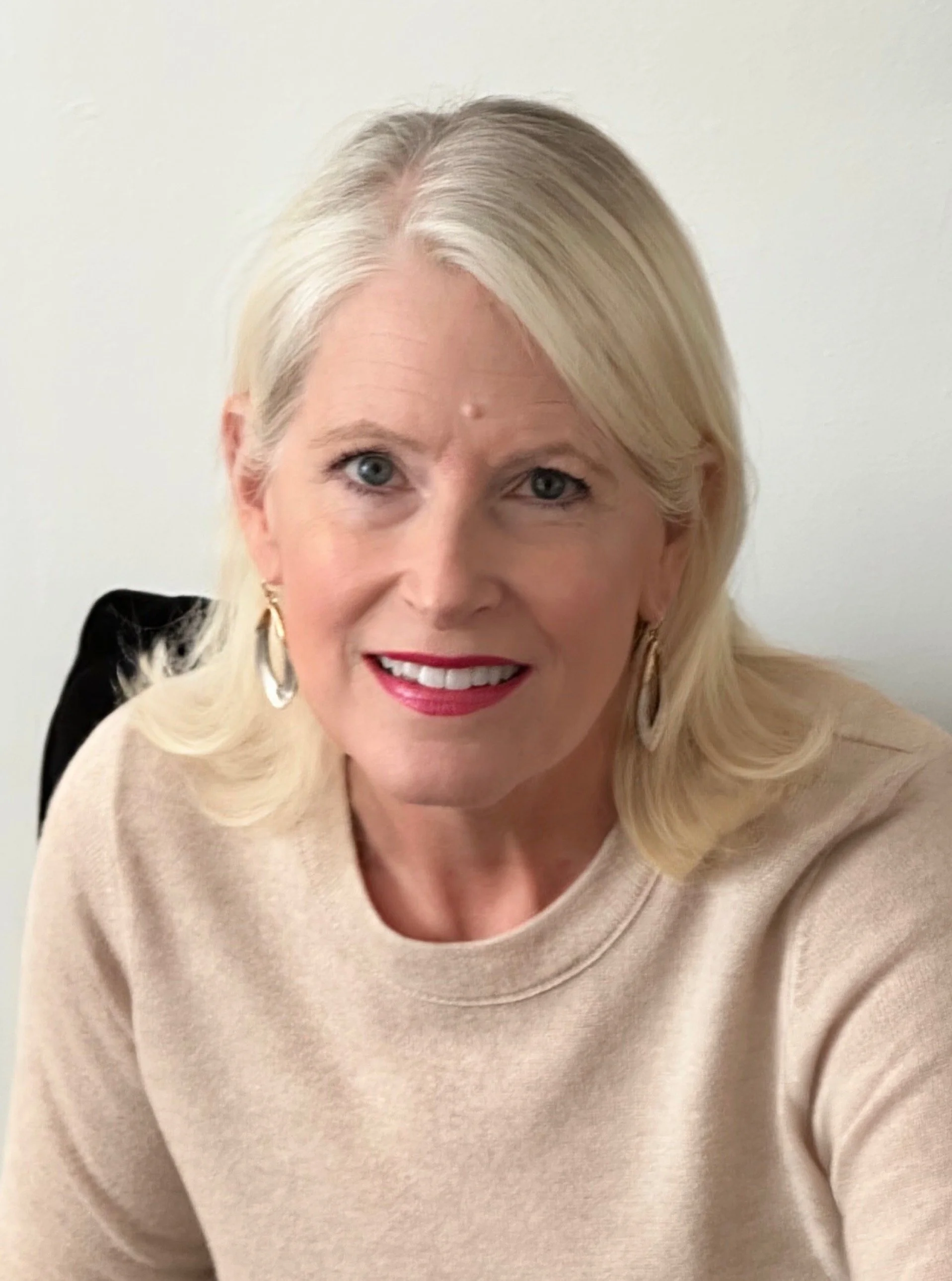 A woman with blonde hair, blue eyes, wearing a beige top and jewelry, smiling against a light wall background.
