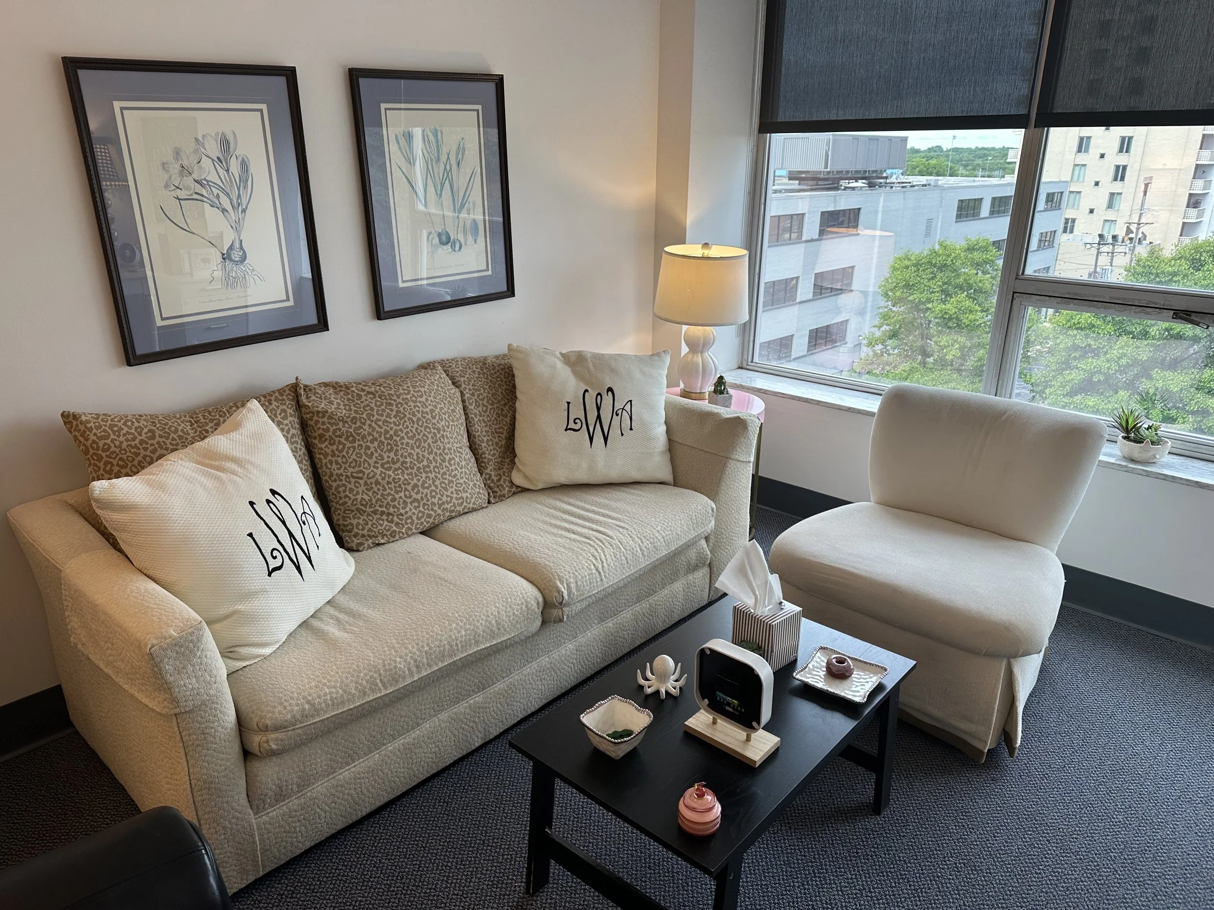 Living room with cream-colored sofa and white armchair near large windows, decorated with framed floral artwork, a table lamp, and various decorative items on a black coffee table.