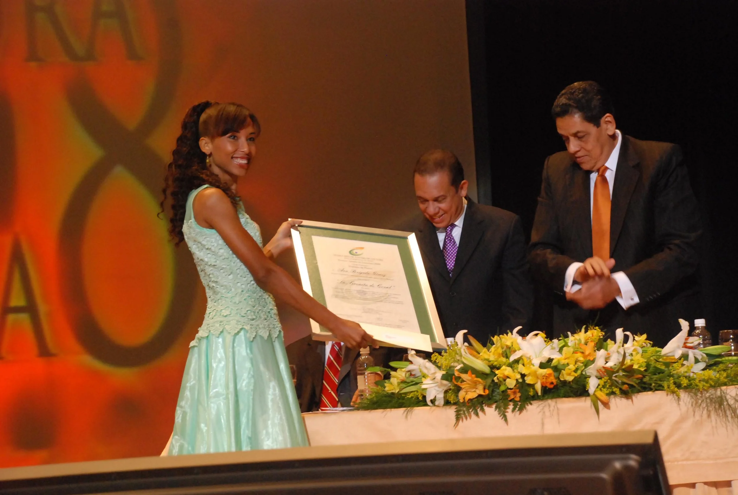 A woman, the author Ana Brigida Gomez, smiling in a light green dress, receives a framed award from a man in a dark suit at a formal event, with two other men watching with a flower-decorated table between them.