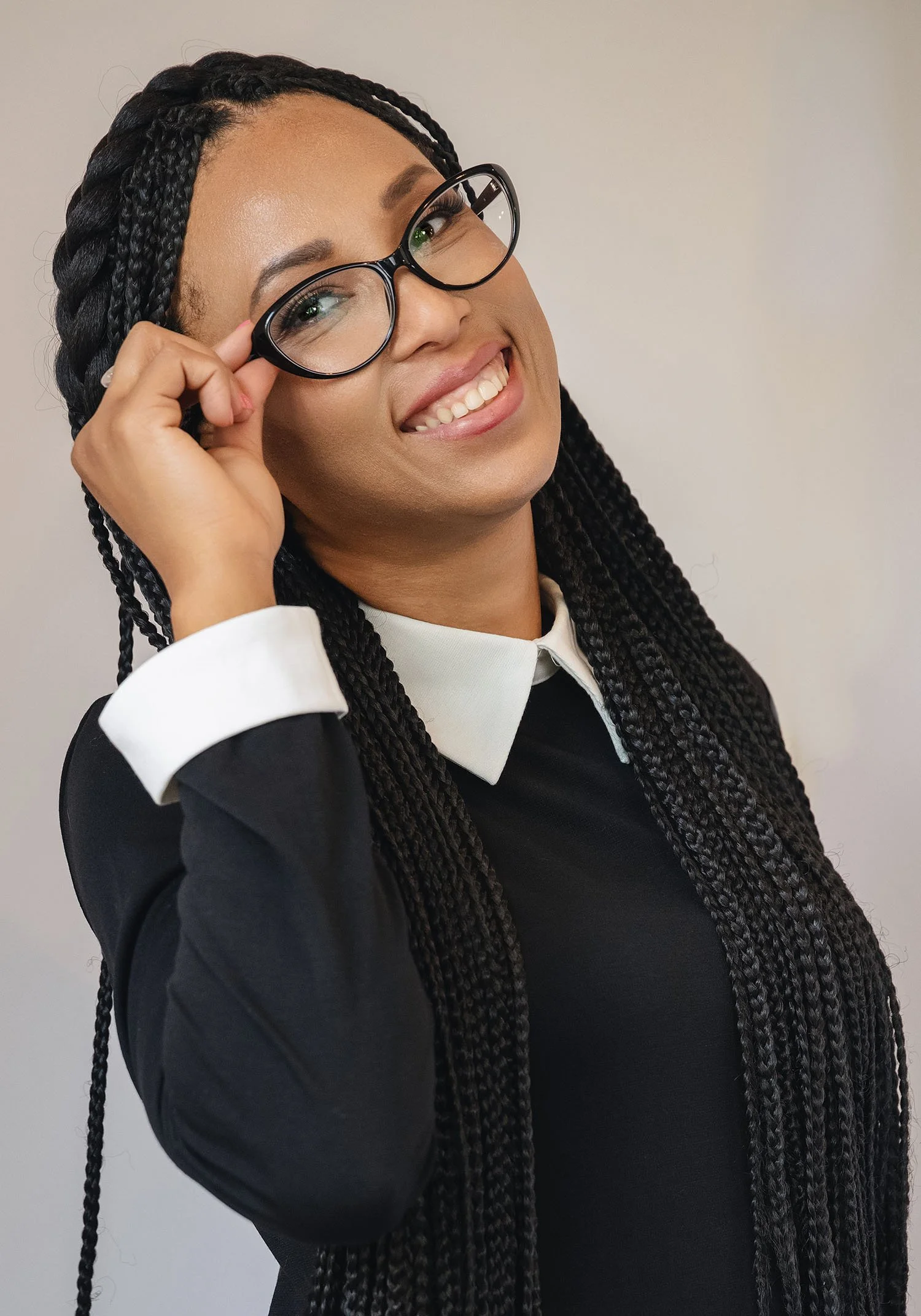 A woman, author Ana Brigida Gomez, with long braided hair dressed in a black jacket, white shirt, smiling, and adjusting her glasses against a plain background.