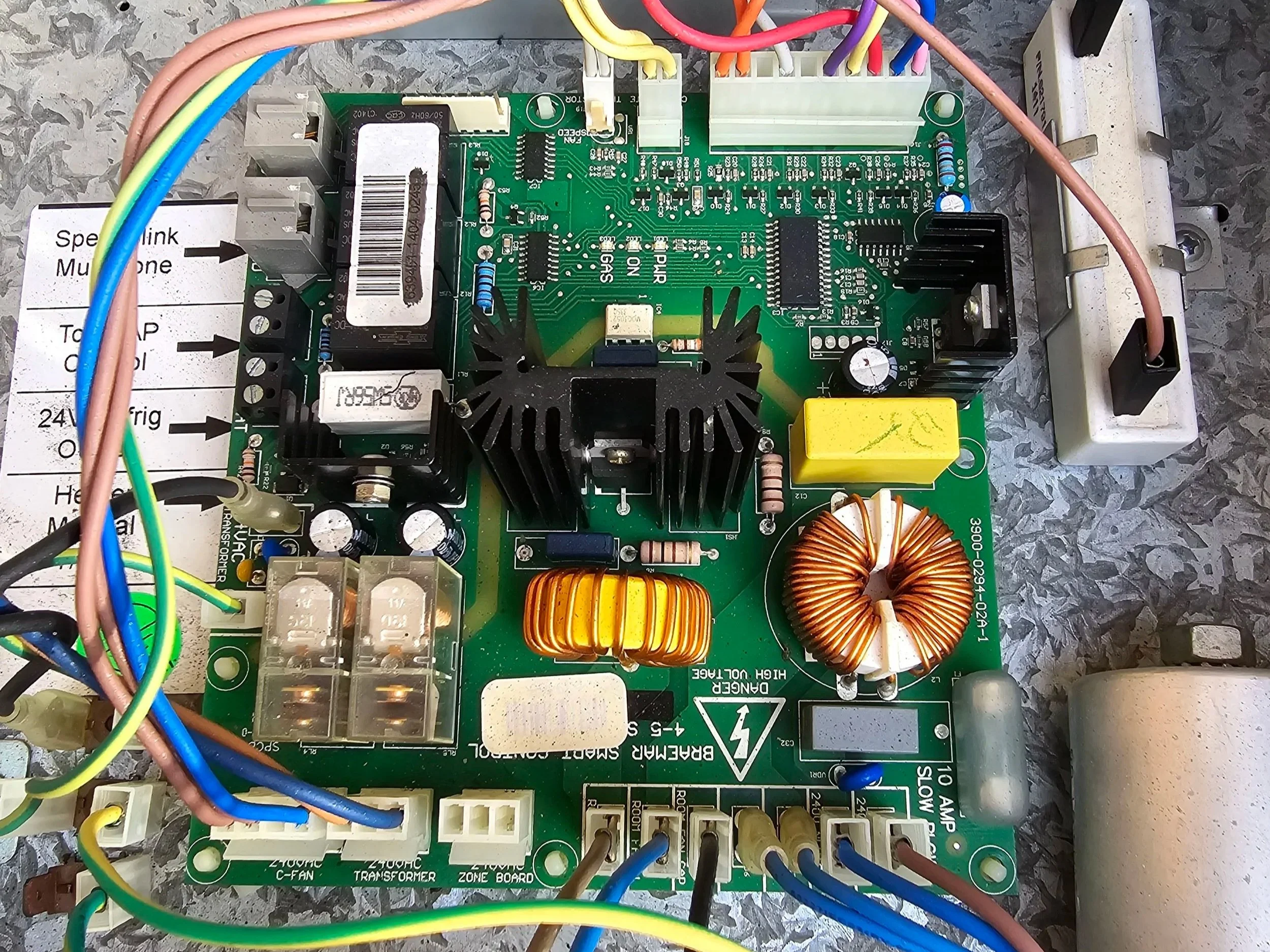Close-up of a green circuit board with various electronic components, yellow and orange coils, blue and pink wires, and a white power strip in the background.