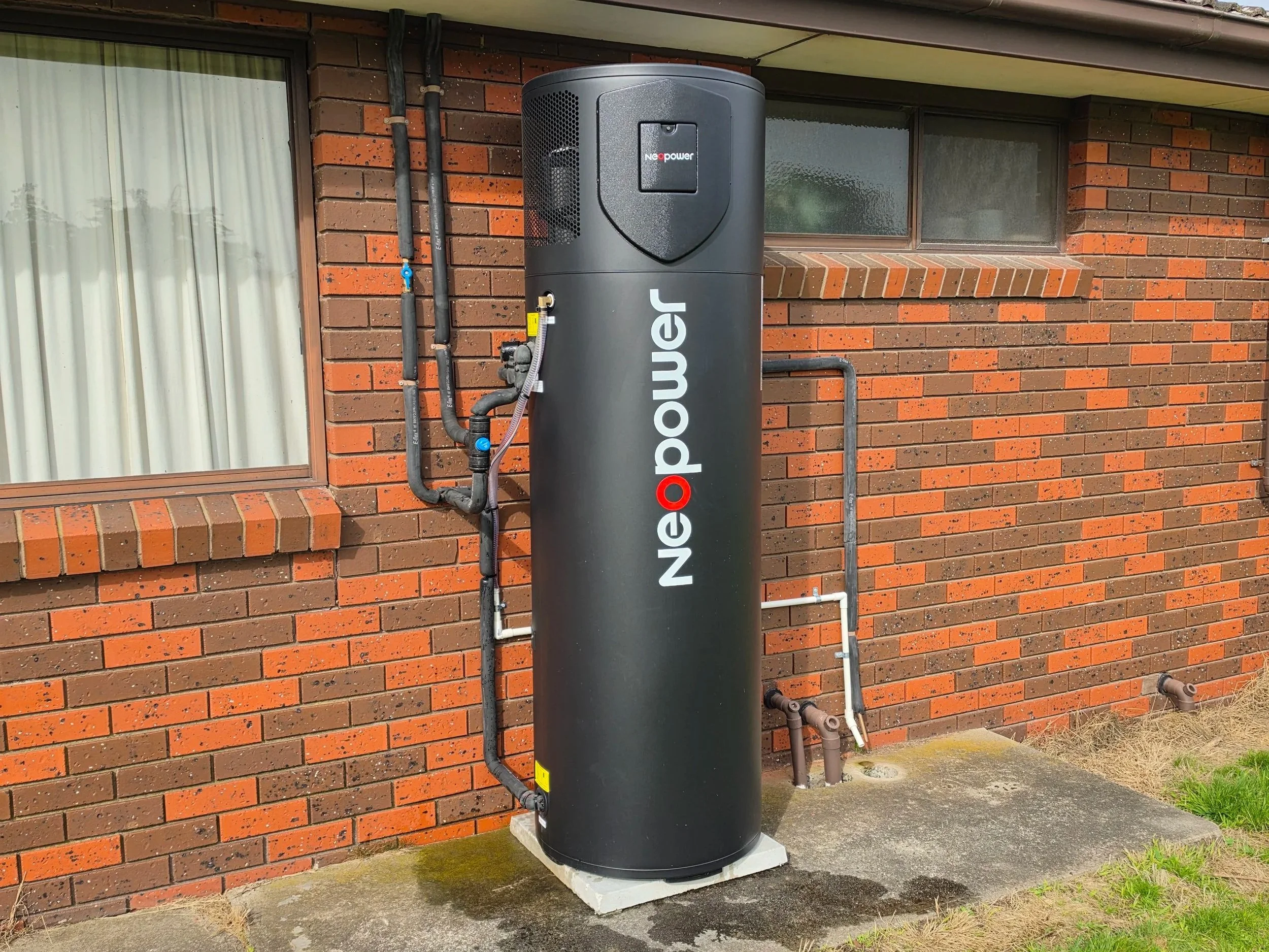 Black Neopower Black Shield heat pump installed outside a brick house with a window and pipes.