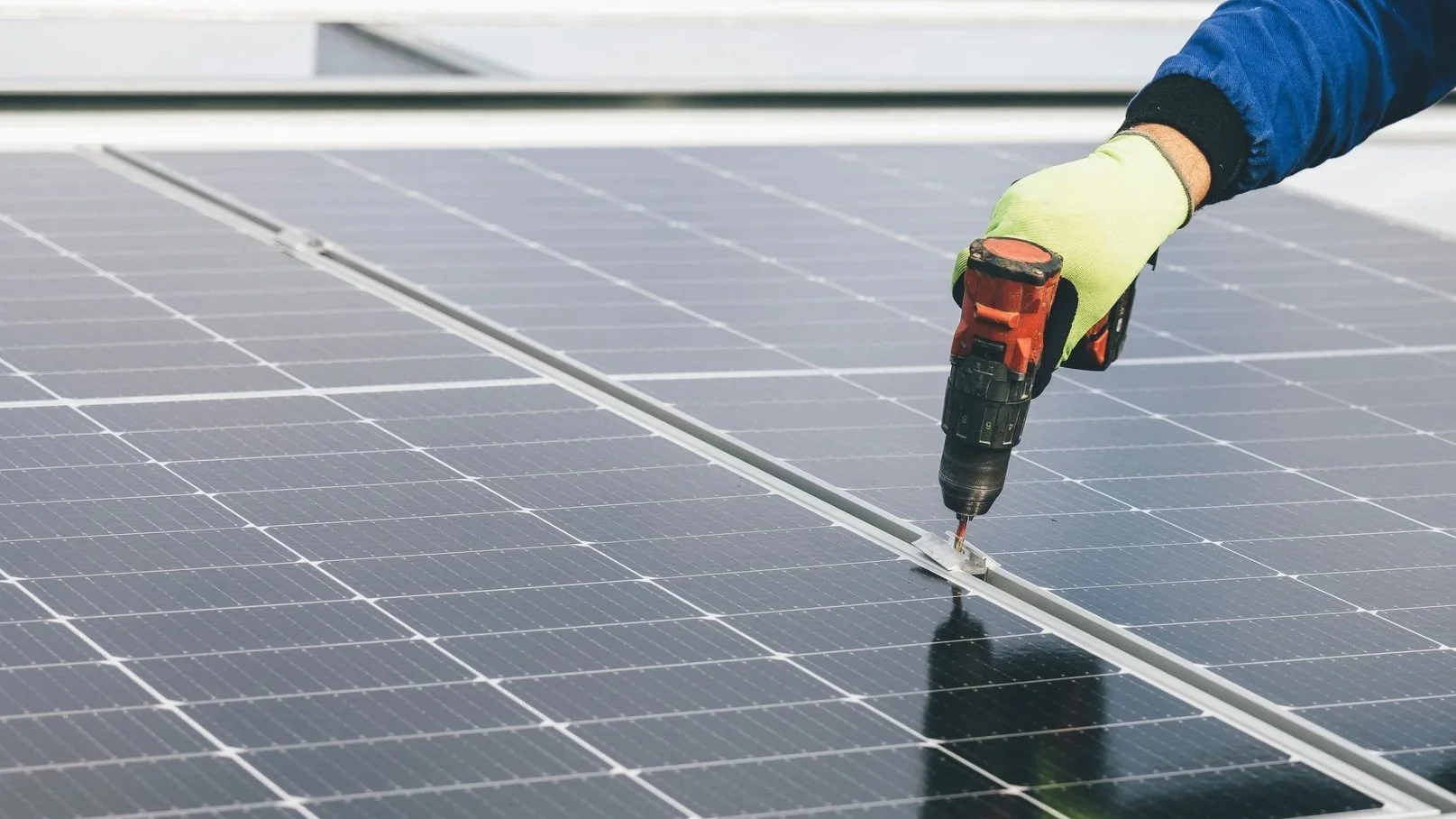 A worker installing or repairing solar panels with a cordless drill, wearing a blue jacket and yellow gloves.