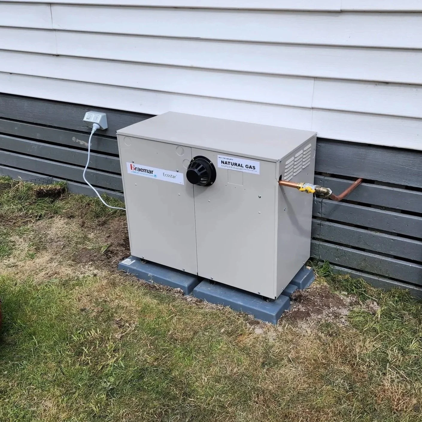 Outdoor natural gas generator unit beside a house with siding, connected to a pipe and electrical outlet.