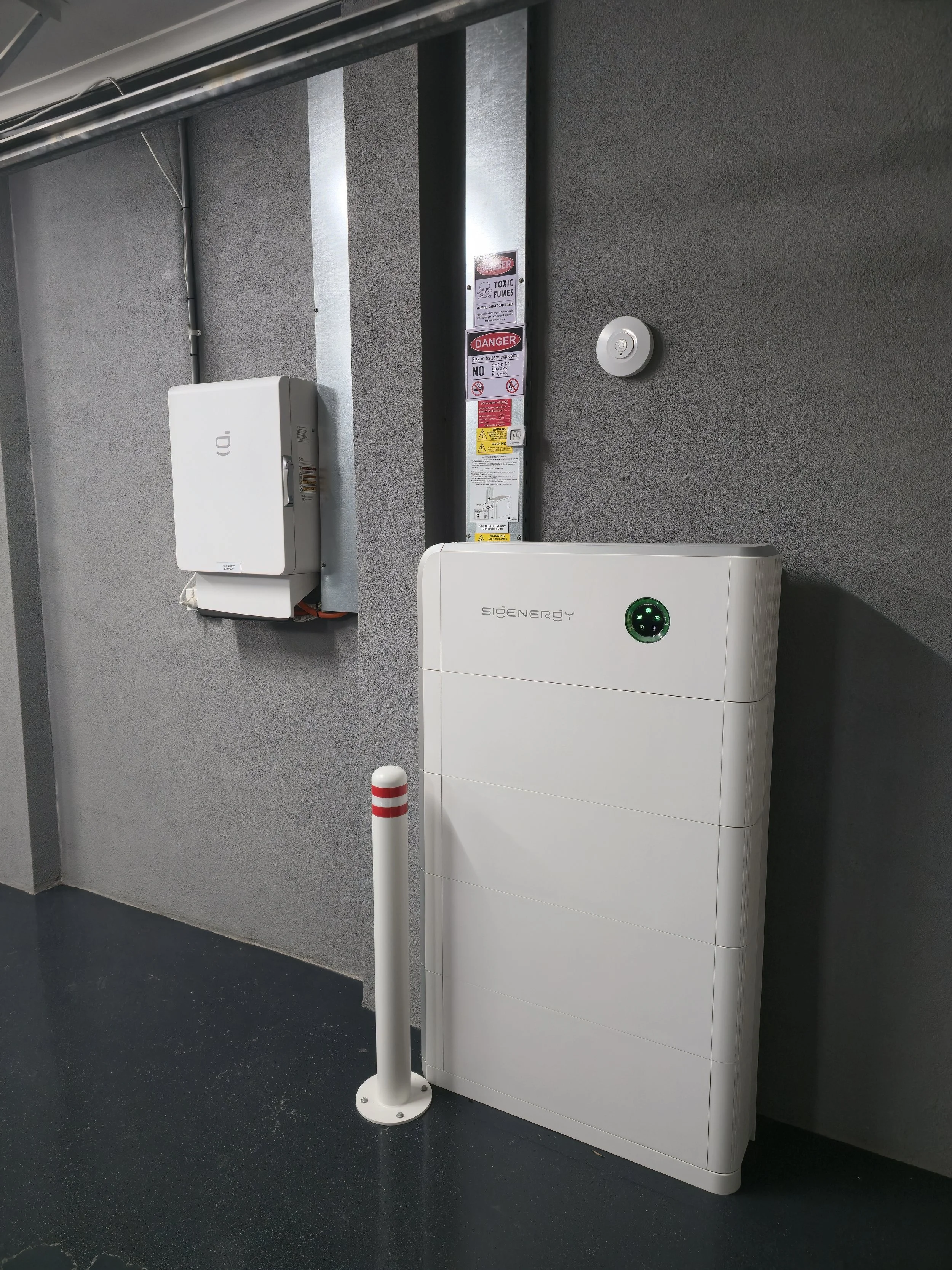 Indoor scene with a Sigenergy Battery Installation, Sigenergy Gateway,  a white pole, security and warning signs, and a dark wall and floor.
