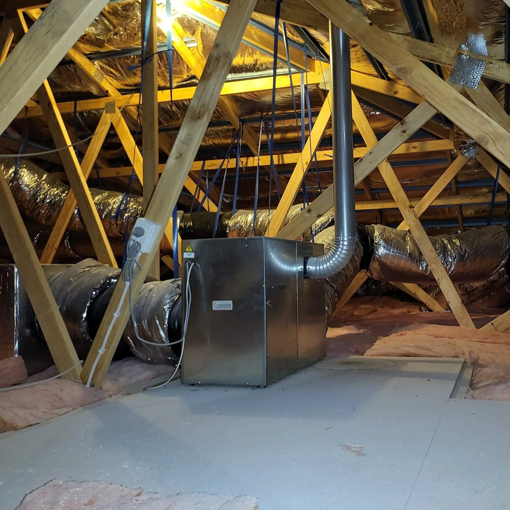 Interior view of an attic showing wooden roof trusses, insulation, a HVAC unit with a connected duct, and some electrical wiring.
