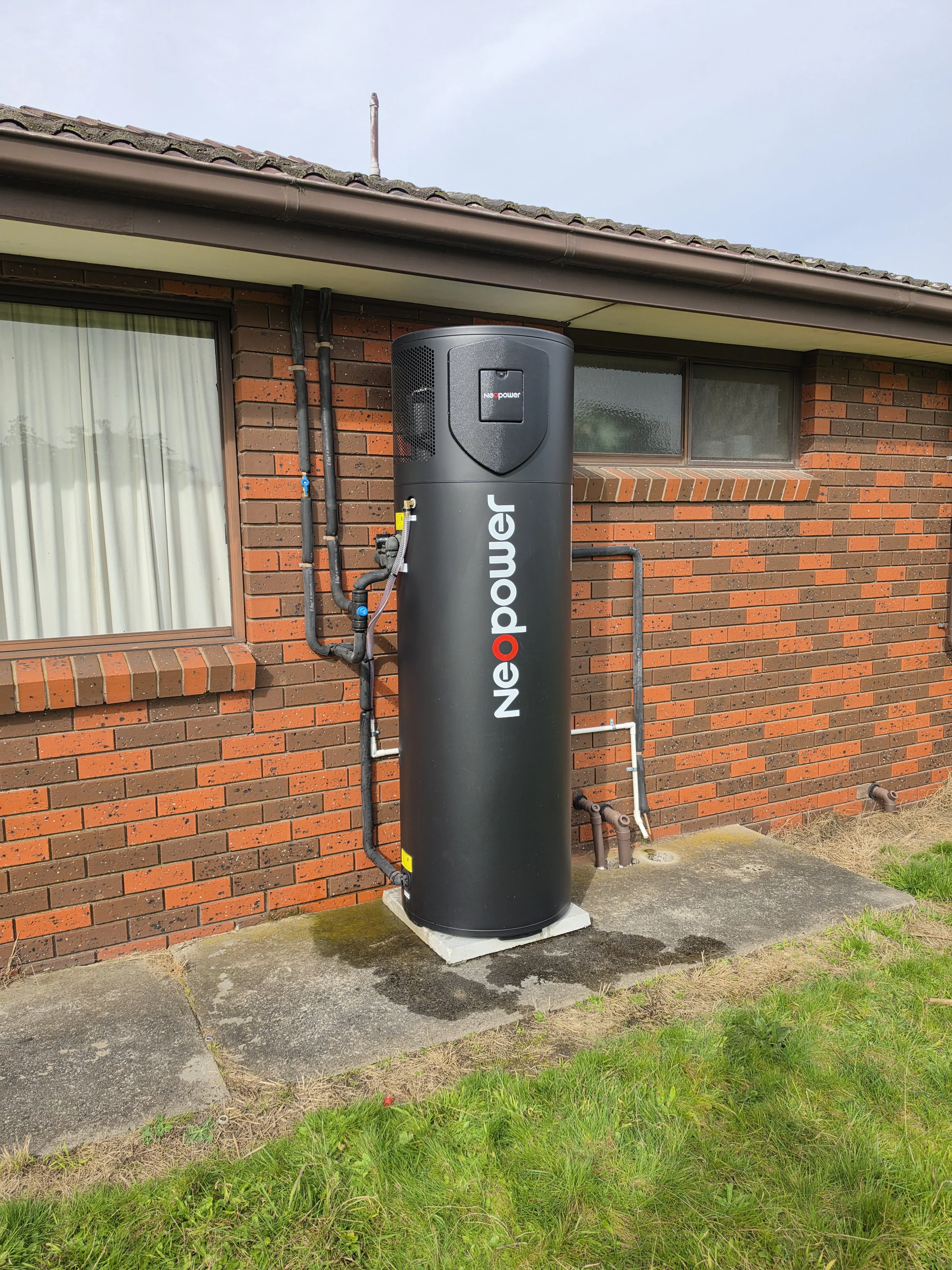 A black tankless water heater system installed outside a brick house, with pipes connected and a small concrete pad, under an overcast sky.