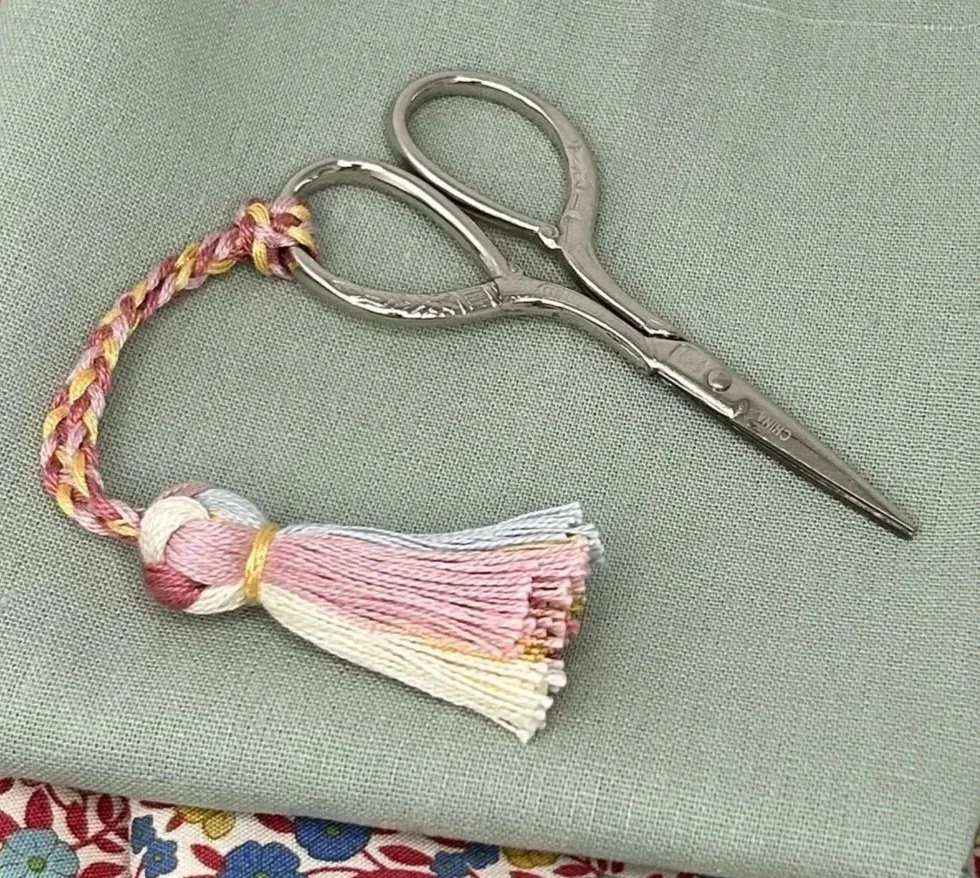 A pair of embroidery scissors with a colorful tassel attached to the handle, resting on a green fabric surface.