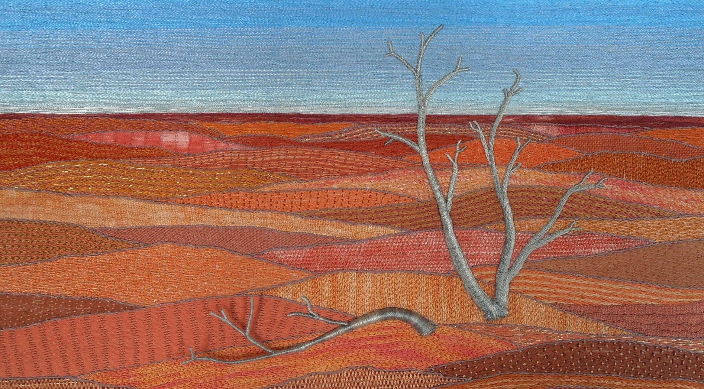 Embroidered panel of red dessert dunes, a big blue sky and a lonely dead tree.