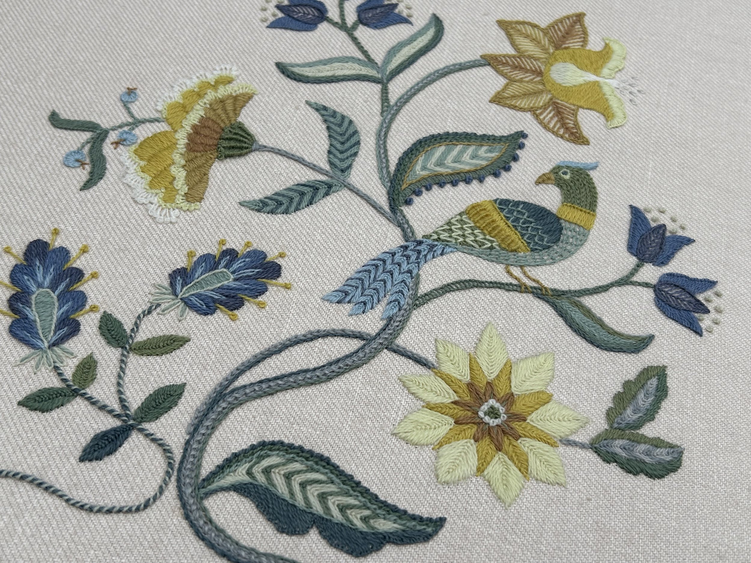 Crewel embroidery. Tree of life design with exotic flowers and a bird stitched in shades of golden yellow, soft greens and deep blue.