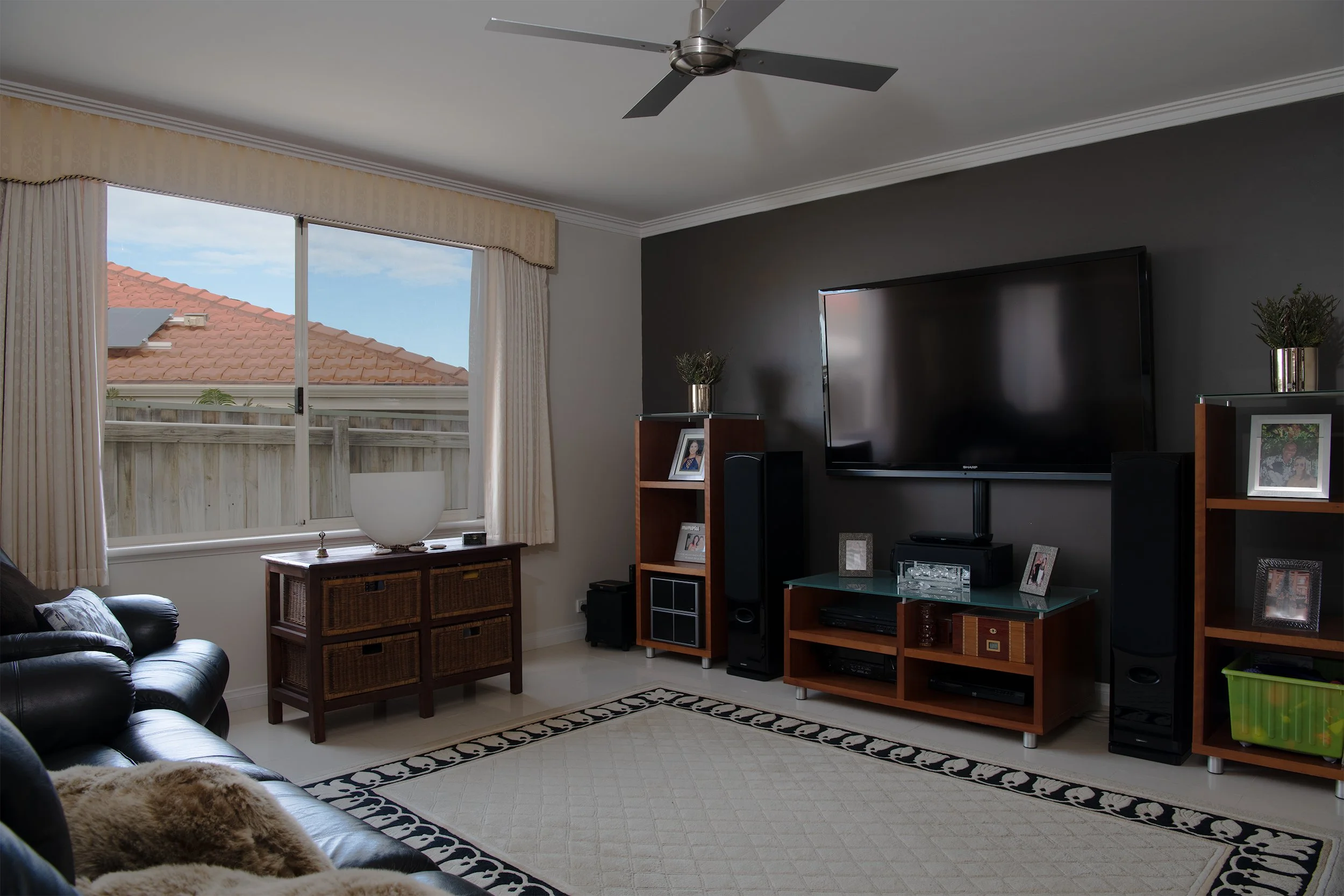 Living room with a large window, ceiling fan, black leather sofa with fur blanket, entertainment center with television, speakers, shelves with photo frames, and a patterned rug.
