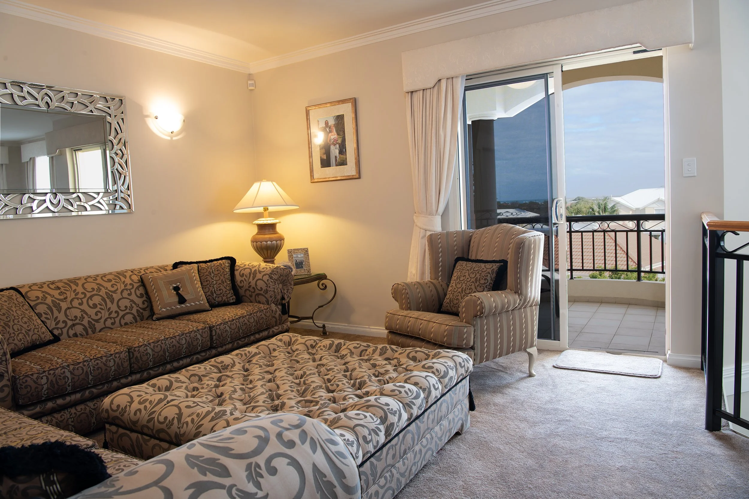 Living room with patterned sofa and armchair, side table with lamp, sliding glass door leading to balcony with ocean view, framed picture on wall, mirror, and beige carpeted floor.