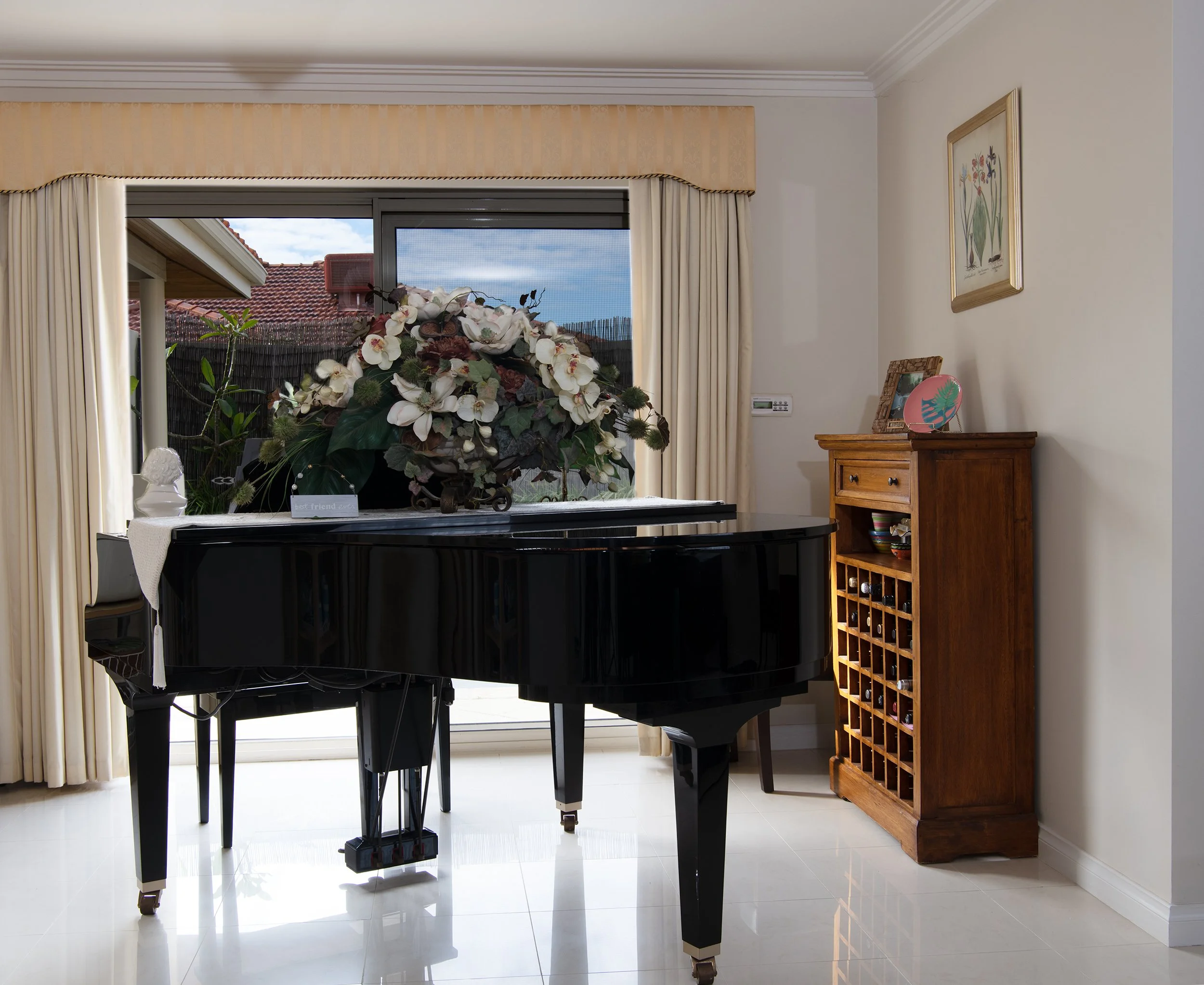 A black grand piano with a floral arrangement on top, located in a bright living room with a large window, beige curtains, and wooden furniture.