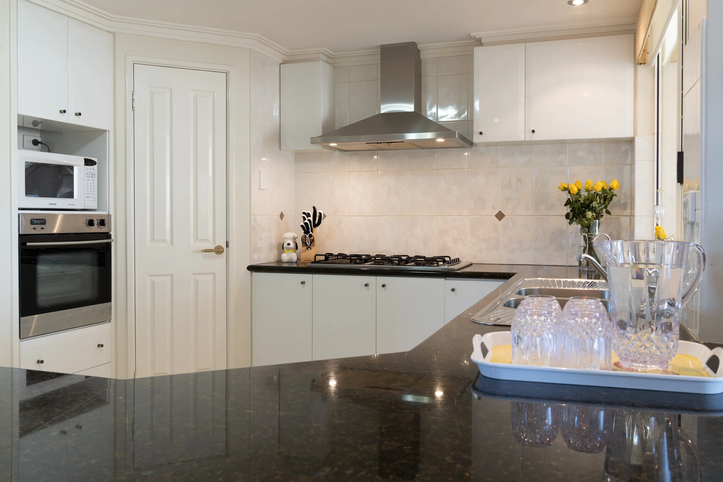 Modern kitchen with white cabinetry, black countertop, integrated microwave and oven, stovetop with stainless steel range hood, vase with yellow flowers, glass pitcher and glasses on tray near double sink, tiled backsplash.