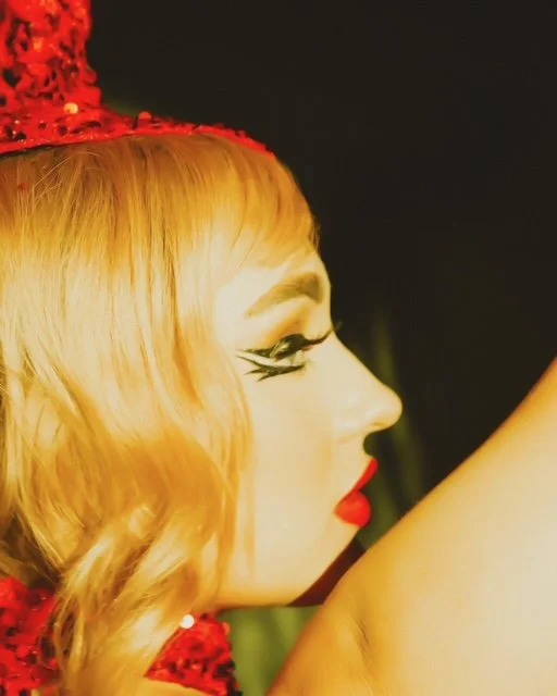 Side profile of a woman with vintage makeup, red lipstick, blonde hair, wearing a red sparkly hat.