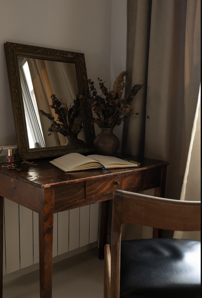 A wooden dresser with an open notebook, a vase with dried flowers, a mirror, and some makeup items, next to beige curtains.