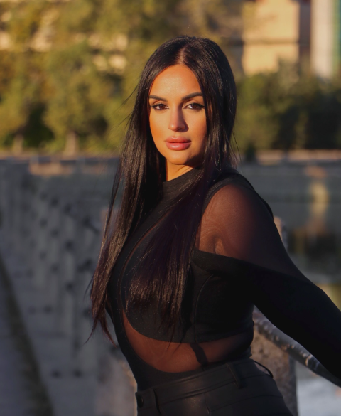 A woman with long dark hair wearing a black sheer top, standing outdoors near a bridge with trees and a building in the background during golden hour sunlight.