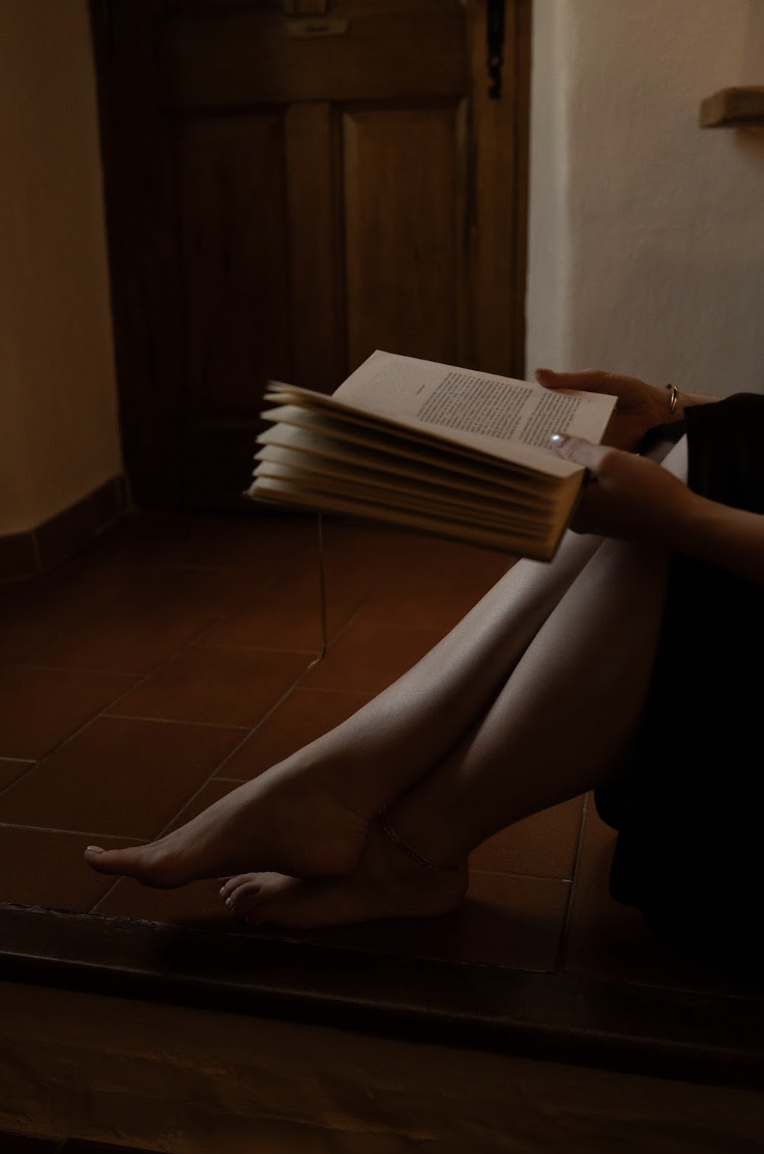 Person sitting on a wooden floor with their legs crossed, reading a large open book. The setting appears to be indoors, with a wooden door in the background.