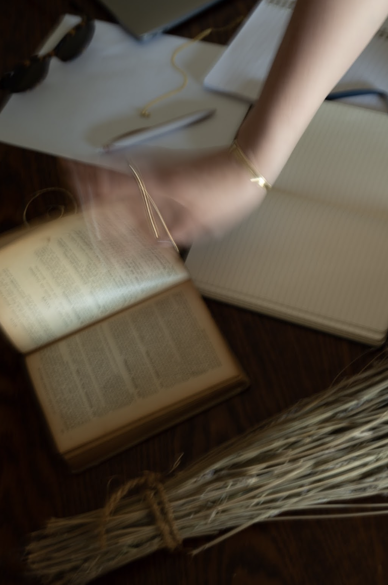 A cluttered desk with an open book, blank notebook, glasses, pen, yellow string, and a bundle of wheat stalks.