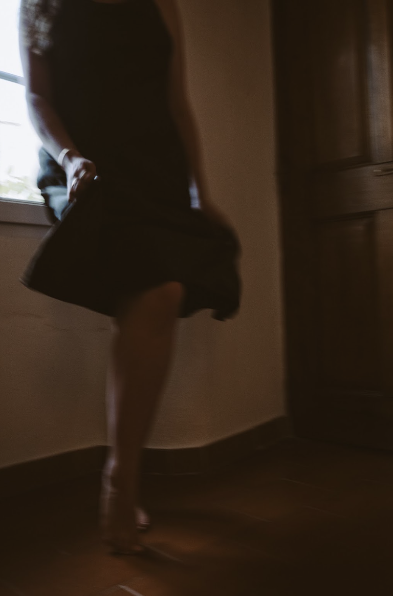 A woman in a dark dress standing on one leg in a room with wooden floors and furniture, with a window nearby.